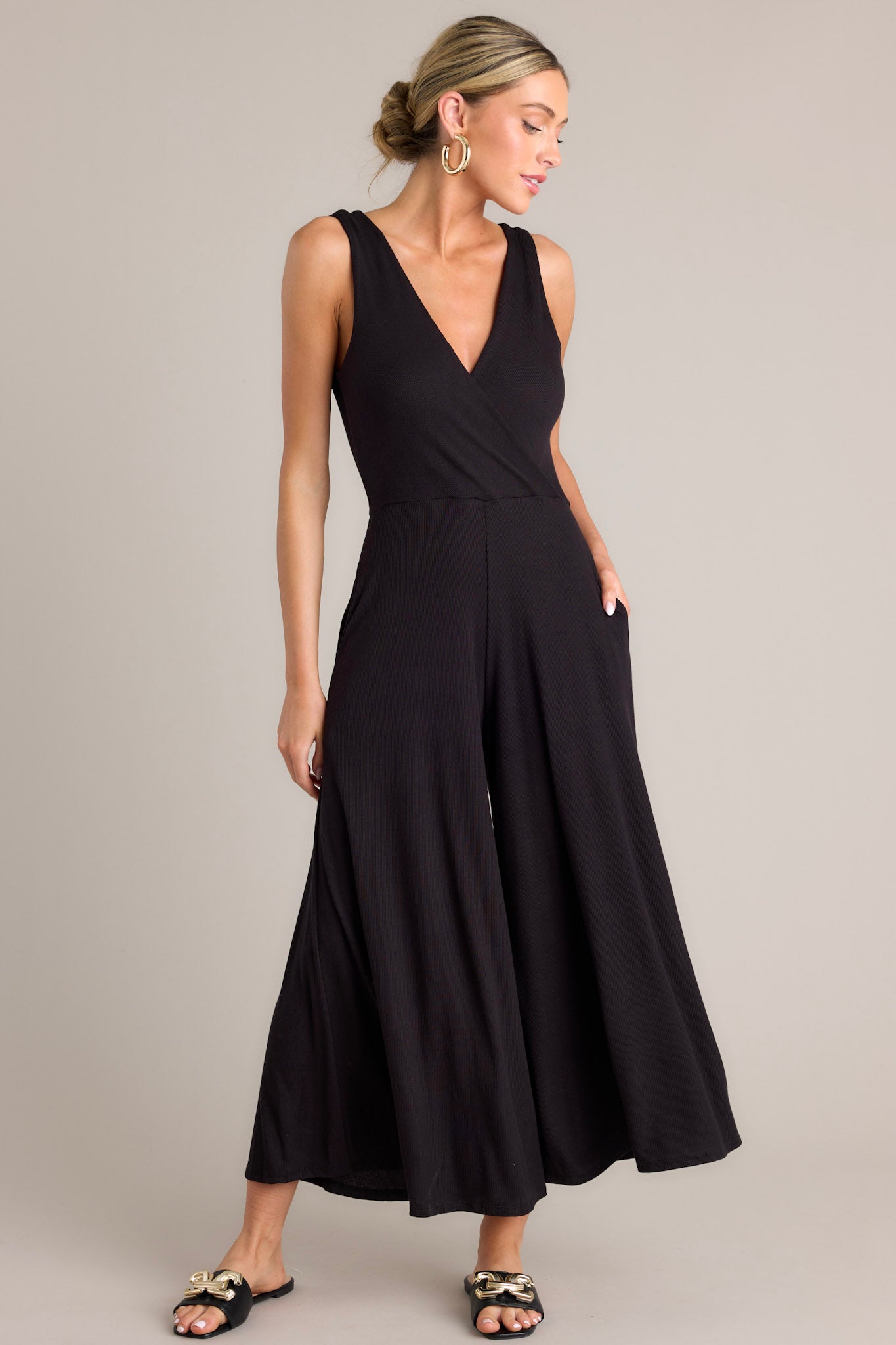 Angled full body view of this black jumpsuit features a v-neckline, a wrap-style bodice, a fitted waist, functional hip pockets, and an extremely wide & flowing leg.