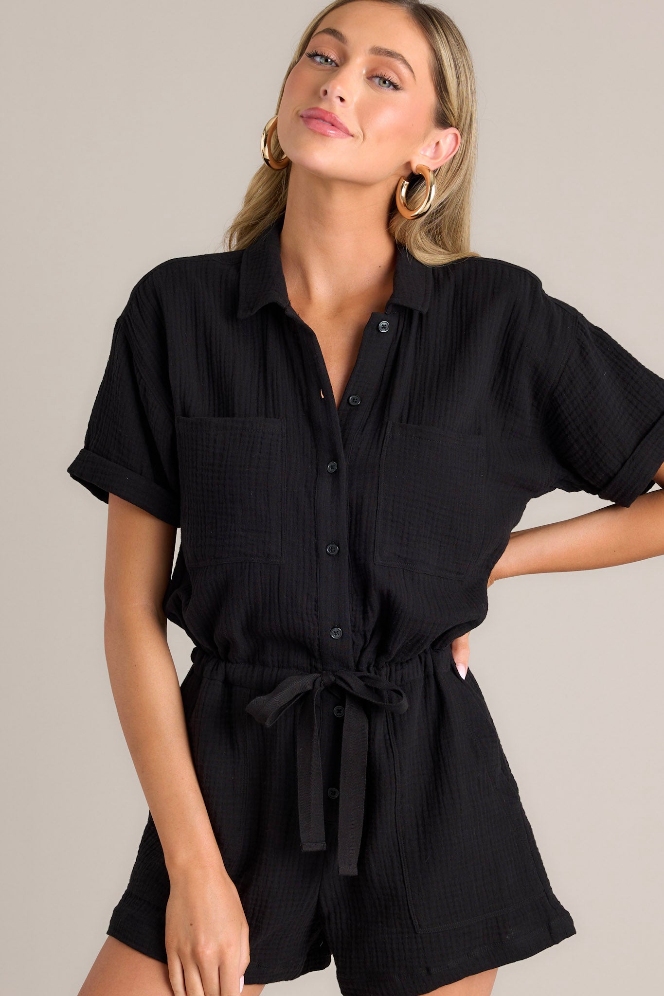 This black romper features a collared neckline, functional breast pockets, a full button front, a self-tie drawstring waist, functional hip pockets, and cuffed short sleeves.