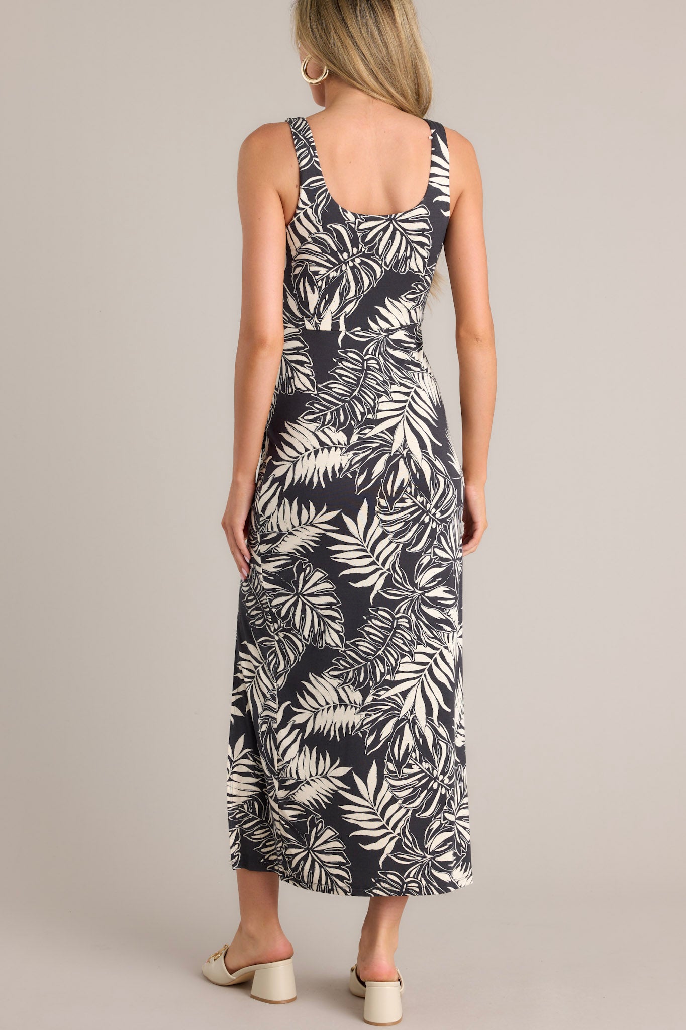 Back view of this charcoal black midi dress that features a scoop neckline, thick straps, an elastic waistband, gathering at the hip, a side slit, and a neutral tropical print.