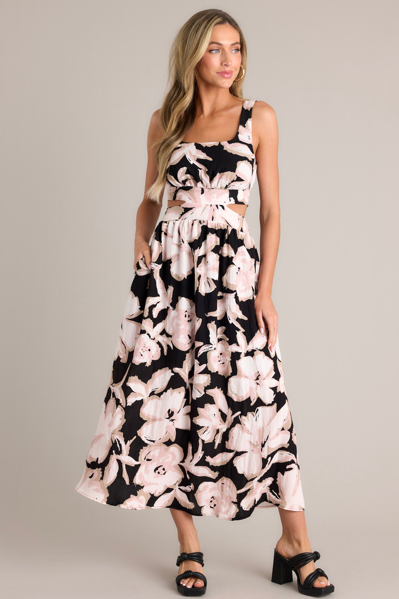 Full body view of this black maxi dress that features a square neckline, thick straps, an elastic back insert, a self-tie back feature, waist cutouts with a smocked back insert, functional hip pockets, and an all over floral pattern.