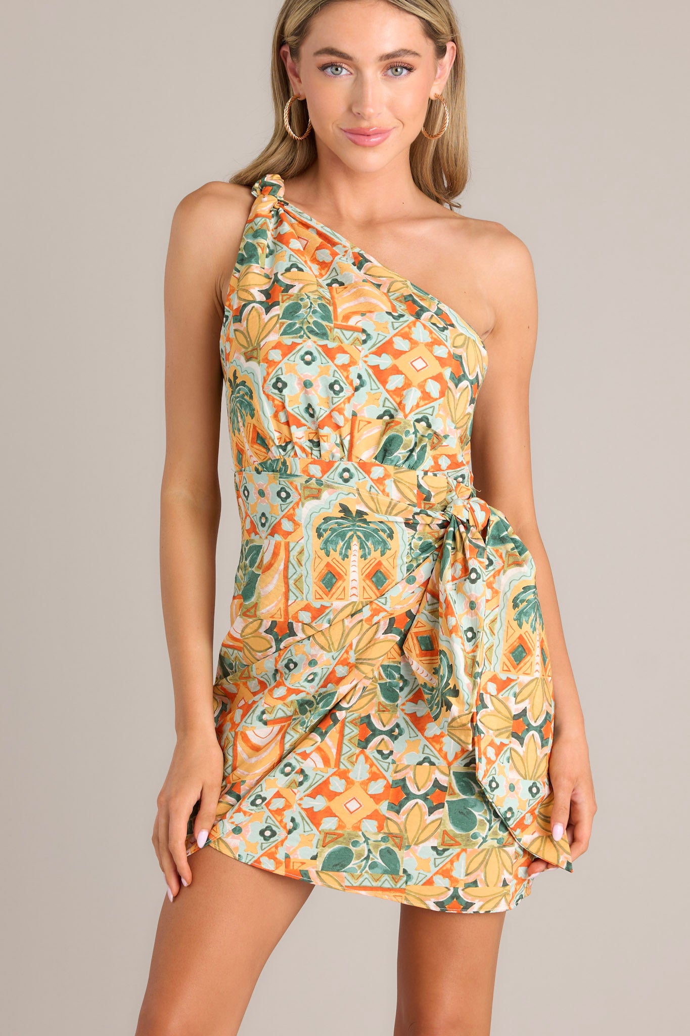 Front view of dress that features an asymmetrical neckline, a knot detail, a smocked back insert, a discrete zipper, a self-tie wrap feature, and a unique eye-catching print.