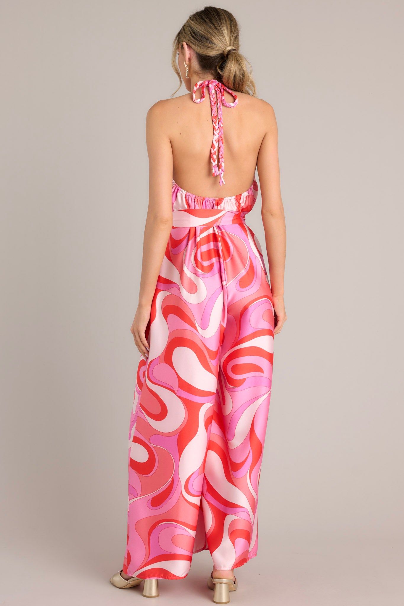 Back view of this jumpsuit that features a high haltered neckline with a braided self tie detail, open back, adjustable belt, pockets at the thigh, and a wide leg.