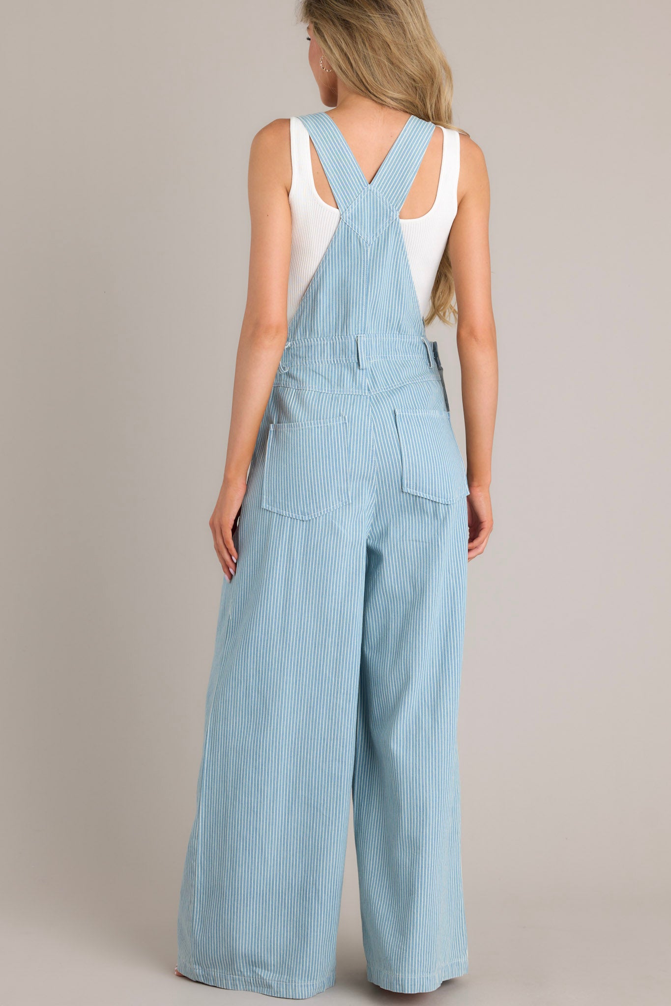 Back view of these overalls that feature a square neckline, adjustable shoulder straps that buckle in the front, a functional center bust pocket, belt loops, functional hip buttons, functional front & back pockets, and a wide leg.