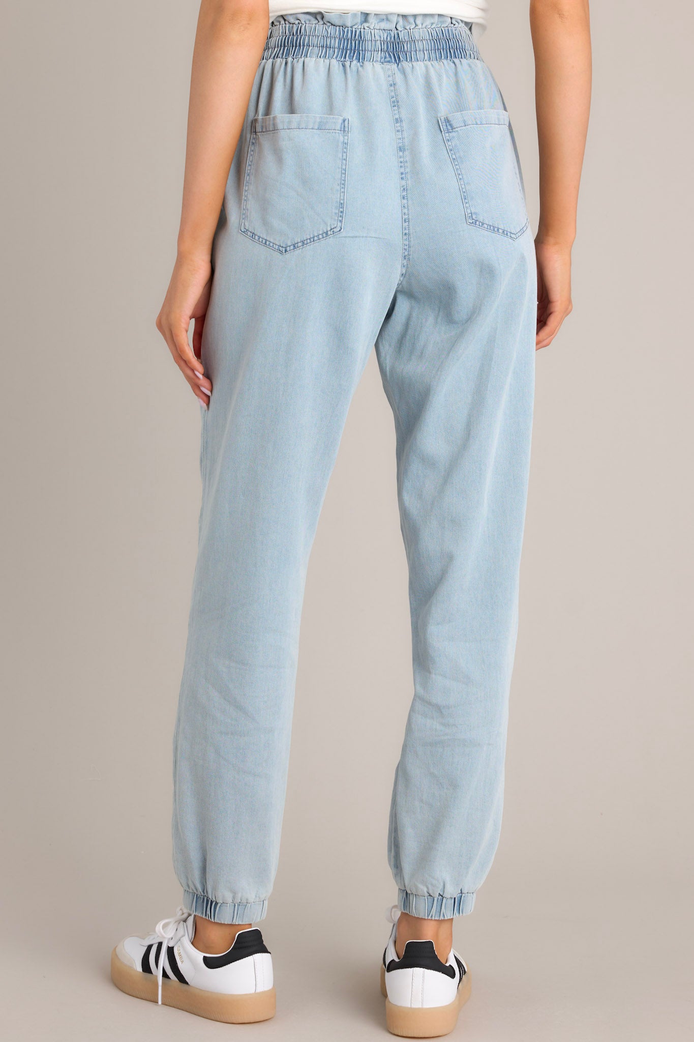 Back view of these light chambray joggers that feature a high waisted design, an elastic paper bag waistband, a self-tie drawstring, functional hip & back pockets, and elastic cuffed ankles.