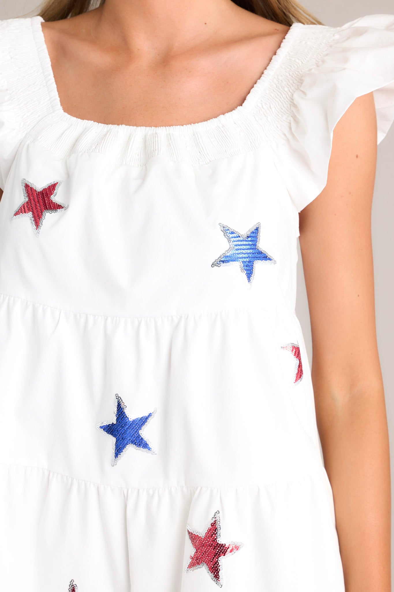 Close up view of this white mini dress that features a smocked square neckline, multicolored sequin stars, a tiered design, and functional hip pockets.