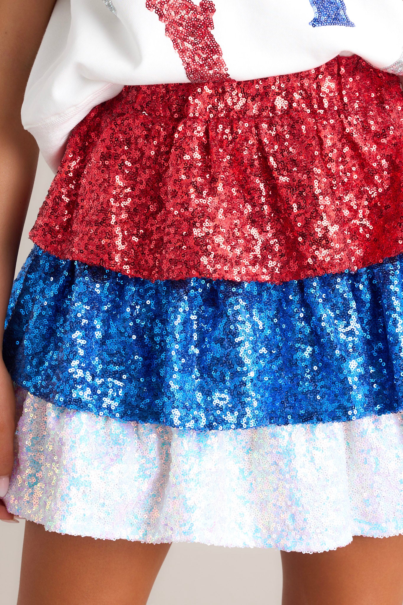 Close up view of this red sequin skirt that features an elastic waistband, sequins throughout, a tiered design, and built in shorts.