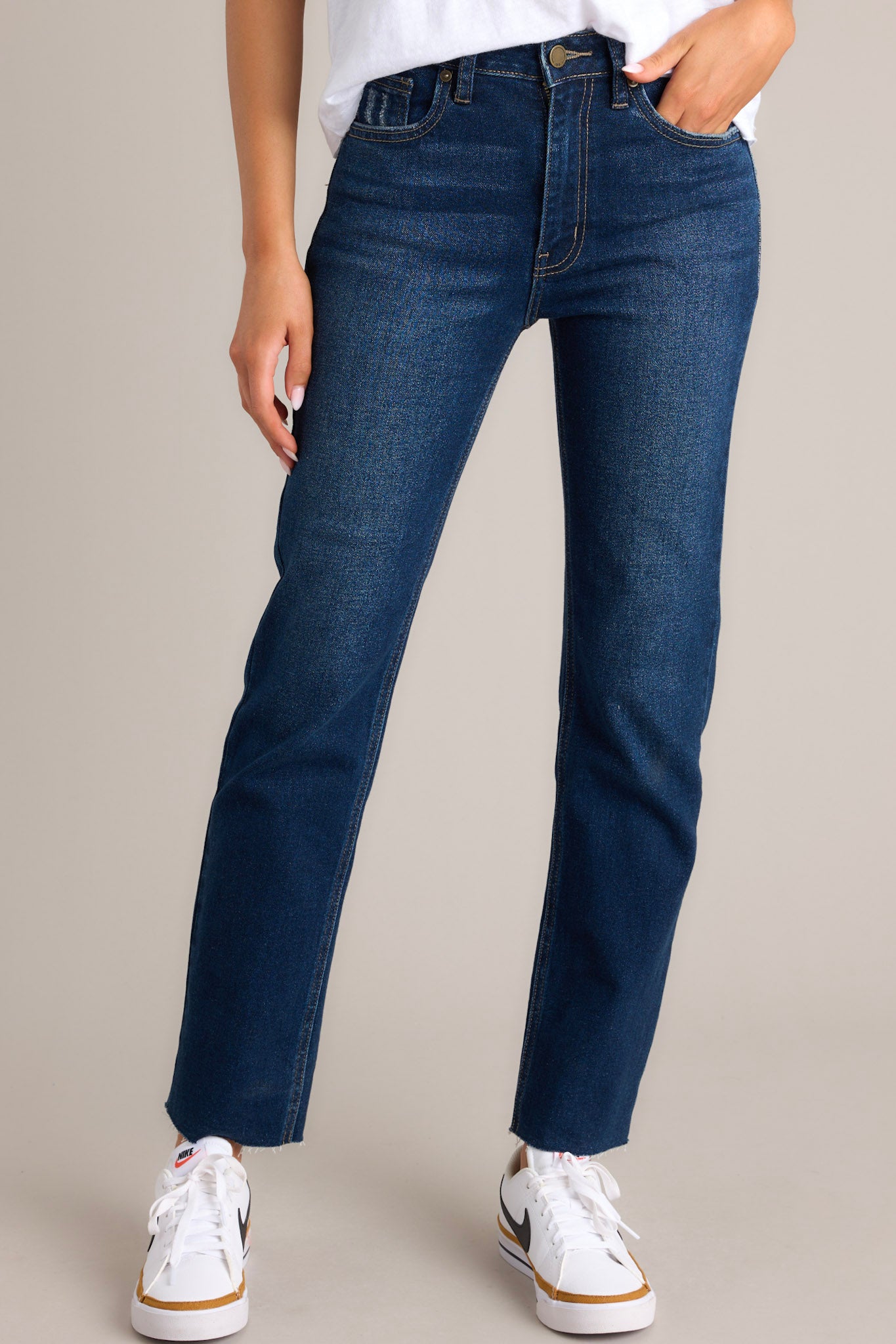 Front angled view of dark wash denim jeans featuring a high-rise waist, scissor cut-off hem, functional pockets, belt loops, and a zipper button closure