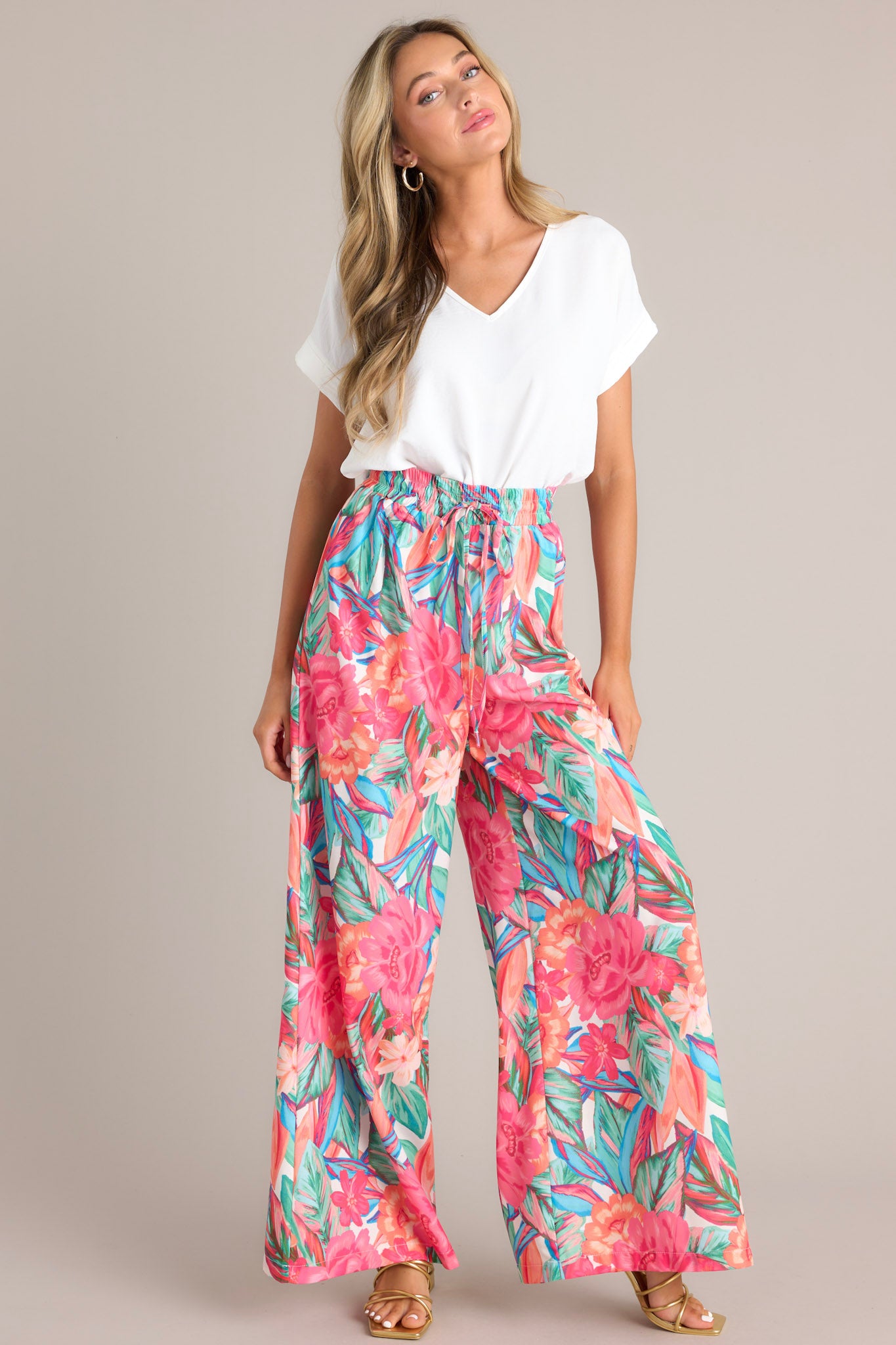Full body angled view of these pink floral pants that feature a high waisted design, an elastic waistband, a self-tie drawstring, a tropical floral print, and a wide leg design.