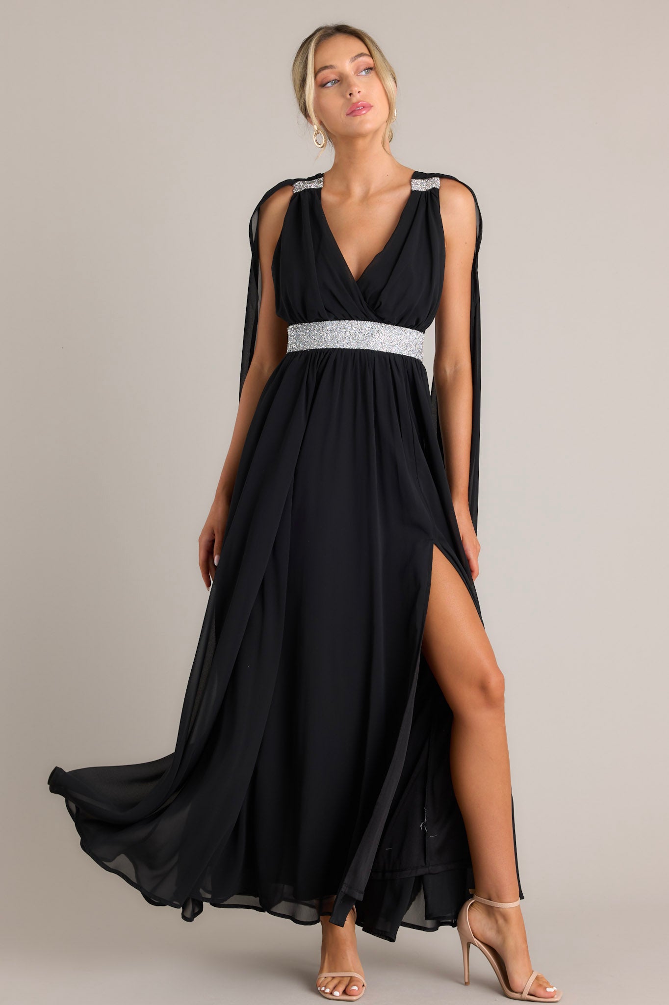 This black dress features a v-neckline, adjustable crisscross straps down the back, rhinestone detailing, fabric trailing from the shoulder to the bottom of the dress, and a slit on the left side of the skirt.
