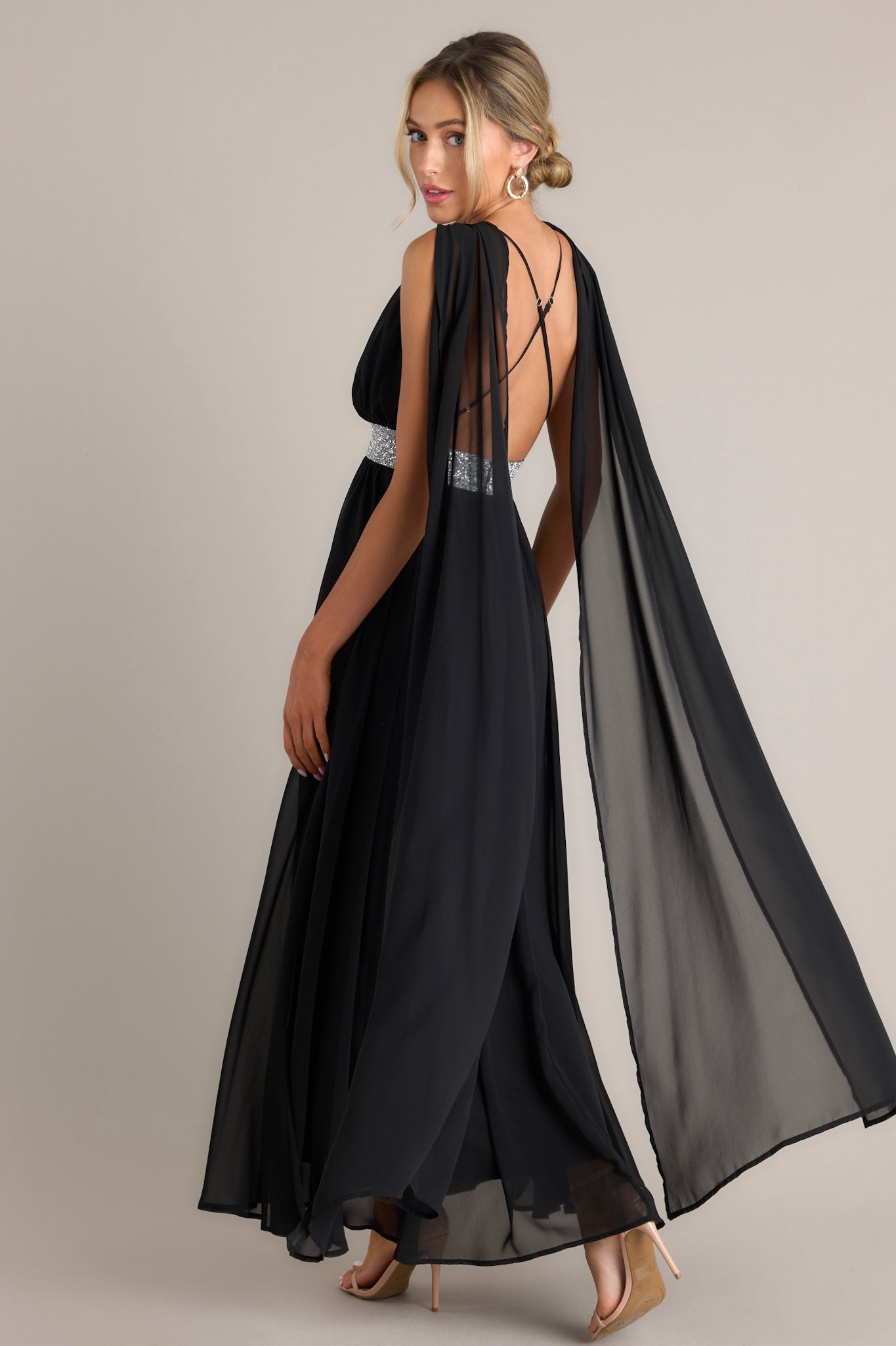 Angled back view of this dress that features a v-neckline, adjustable crisscross straps down the back, rhinestone detailing, fabric trailing from the shoulder to the bottom of the dress, and a slit on the left side of the skirt.