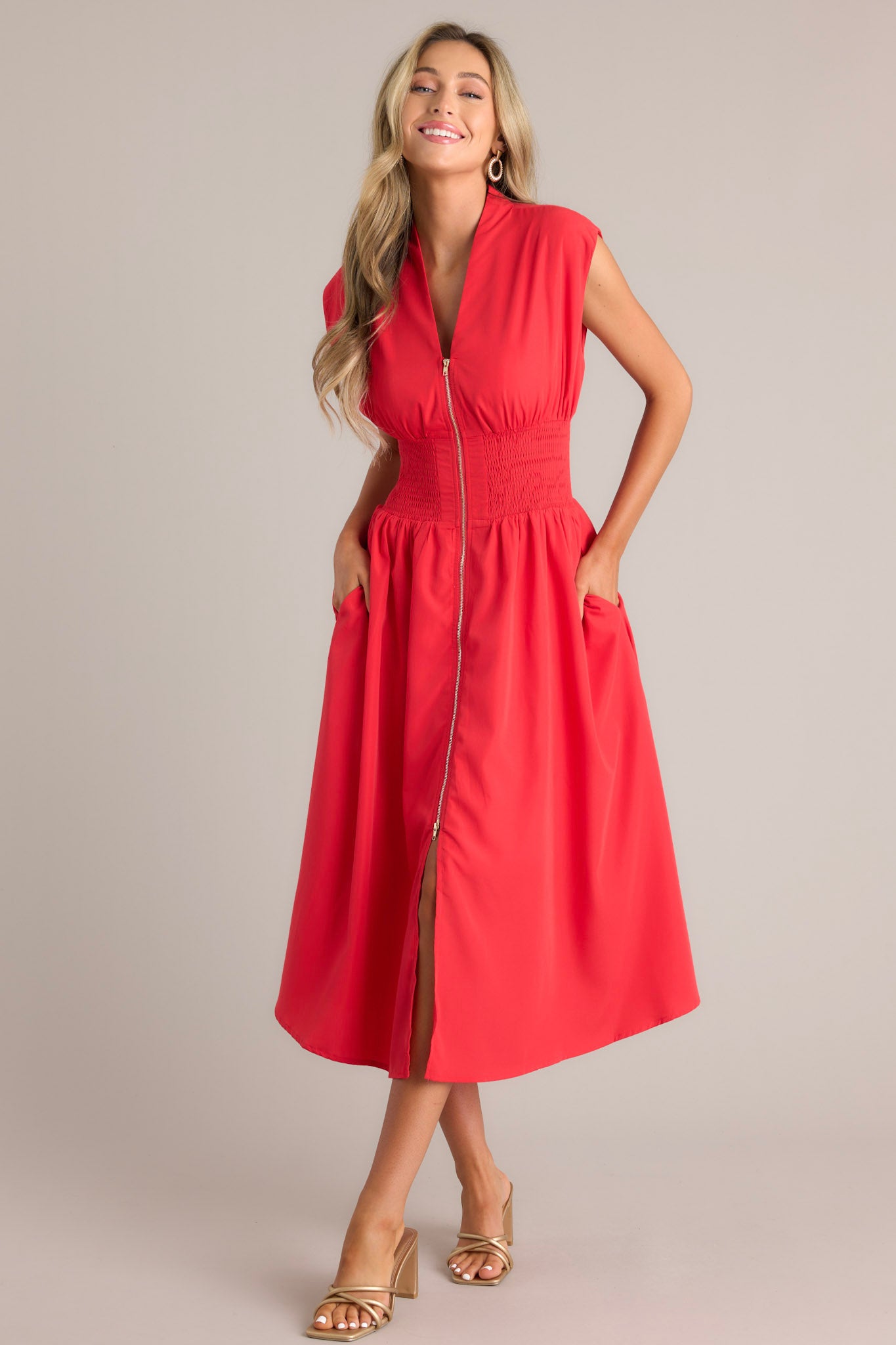 This red midi dress features a v-neckline, padded shoulders, a full zipper front, a fully smocked waist, functional hip pockets, and a front slit.