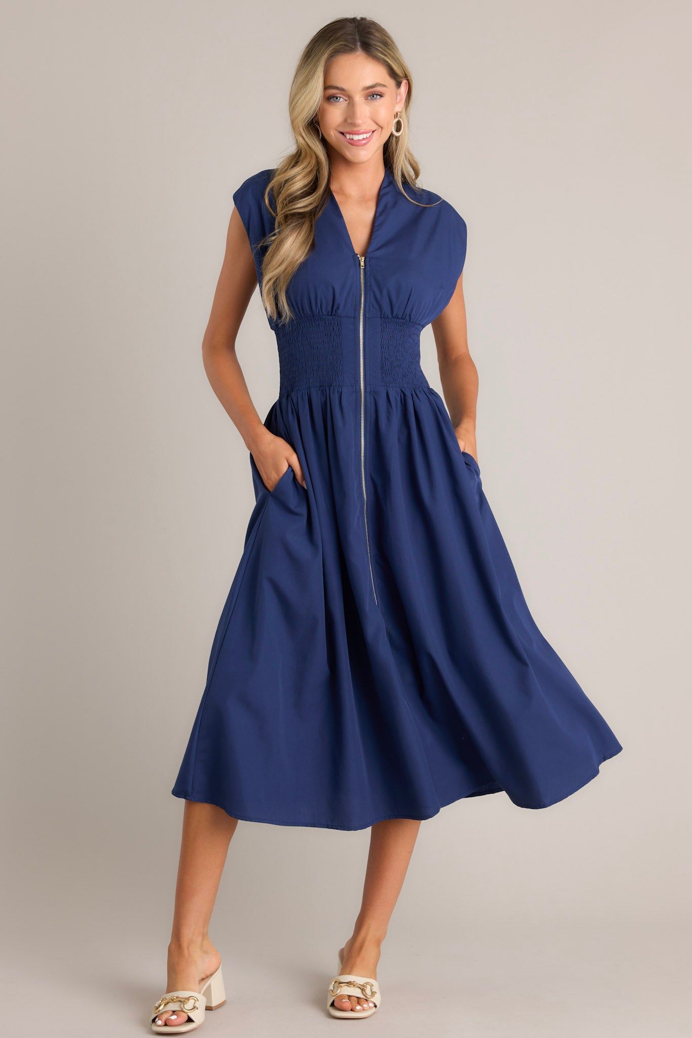 Full body view of a navy blue dress with a V-neckline, cap sleeves, a zippered front, and a cinched waist with shirred detailing, featuring a flowing skirt and side pockets.