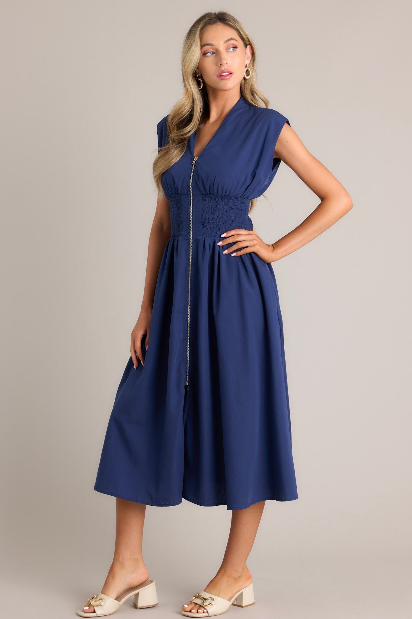 Side view of a navy blue dress with a V-neckline, cap sleeves, a zippered front, and a cinched waist with shirred detailing, featuring a flowing midi-length skirt, highlighting its elegant and stylish design.