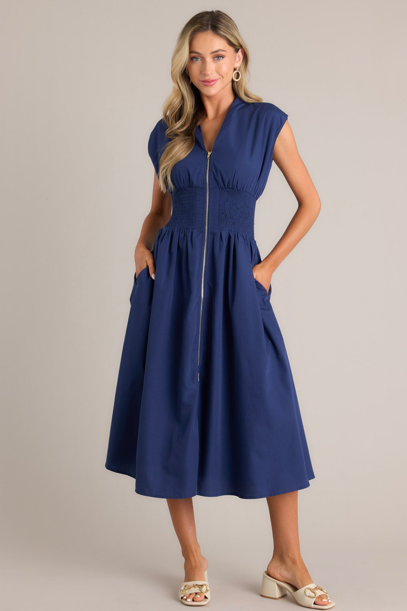 Front view of a navy blue dress with a V-neckline, cap sleeves, a zippered front, and a cinched waist with shirred detailing, featuring a flowing midi-length skirt, highlighting its elegant and stylish design.