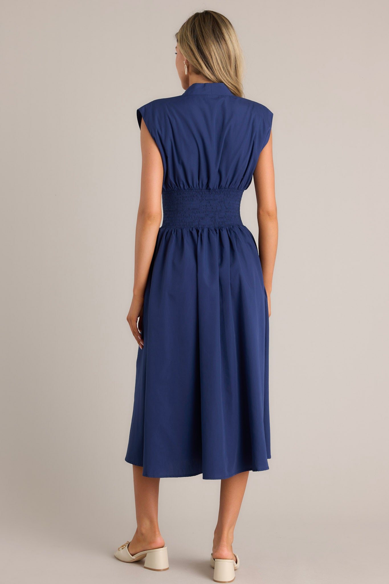 Back view of a navy blue dress with a V-neckline, cap sleeves, a zippered front, and a cinched waist with shirred detailing, featuring a flowing midi-length skirt, highlighting its elegant and stylish design.
