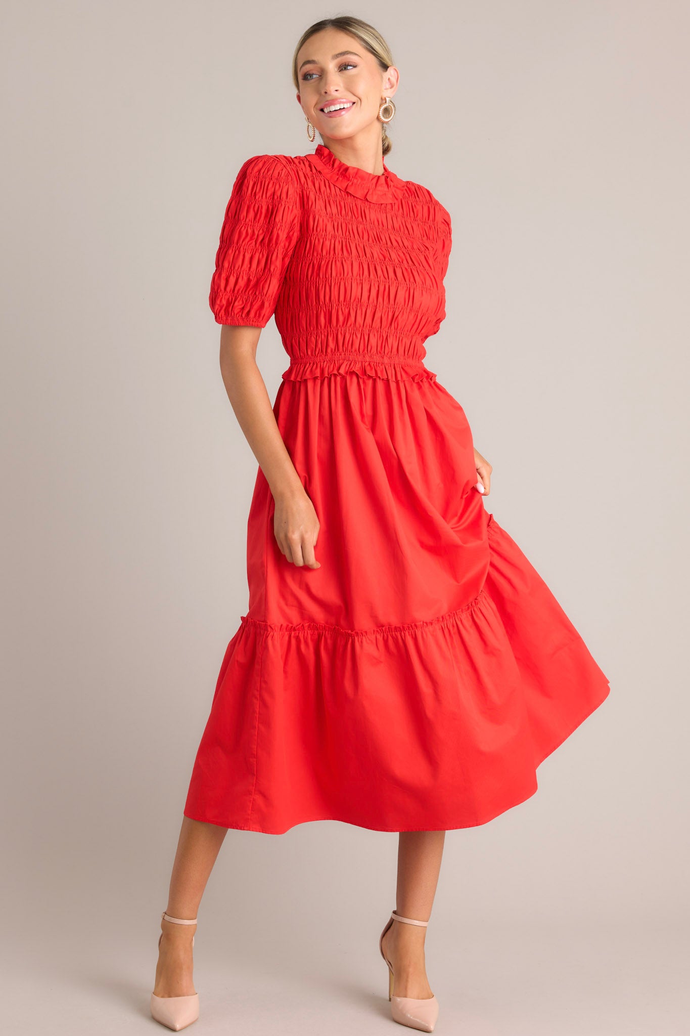Angled front view of this red midi dress that features high ruffled neckline, a keyhole with button closure, a fully smocked bodice, a tiered design, a fully smocked elastic cuffed half sleeves.