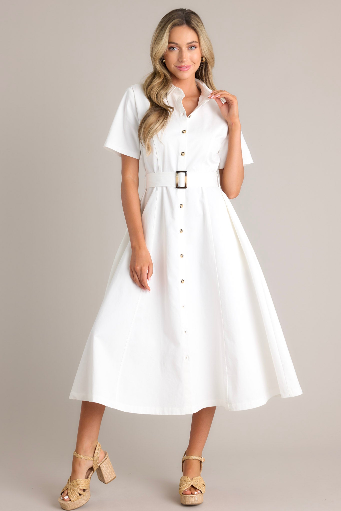 Angled front view of this white midi dress that features a collared neckline, a full button front, a functional pull-through belt, functional hip pockets, and short sleeves.