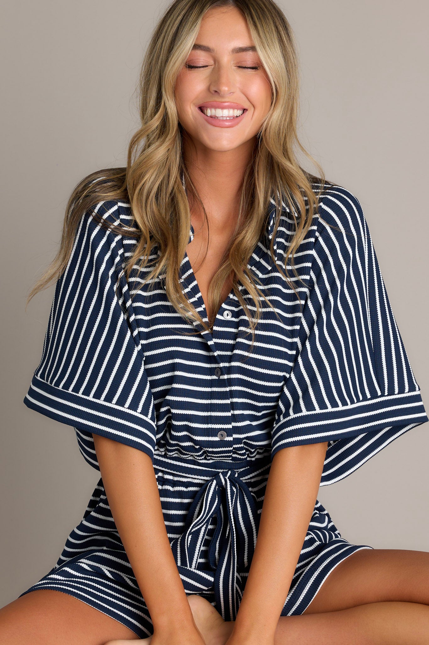 Seated front view of this Navy blue and white striped romper with a front button closure and waist tie.