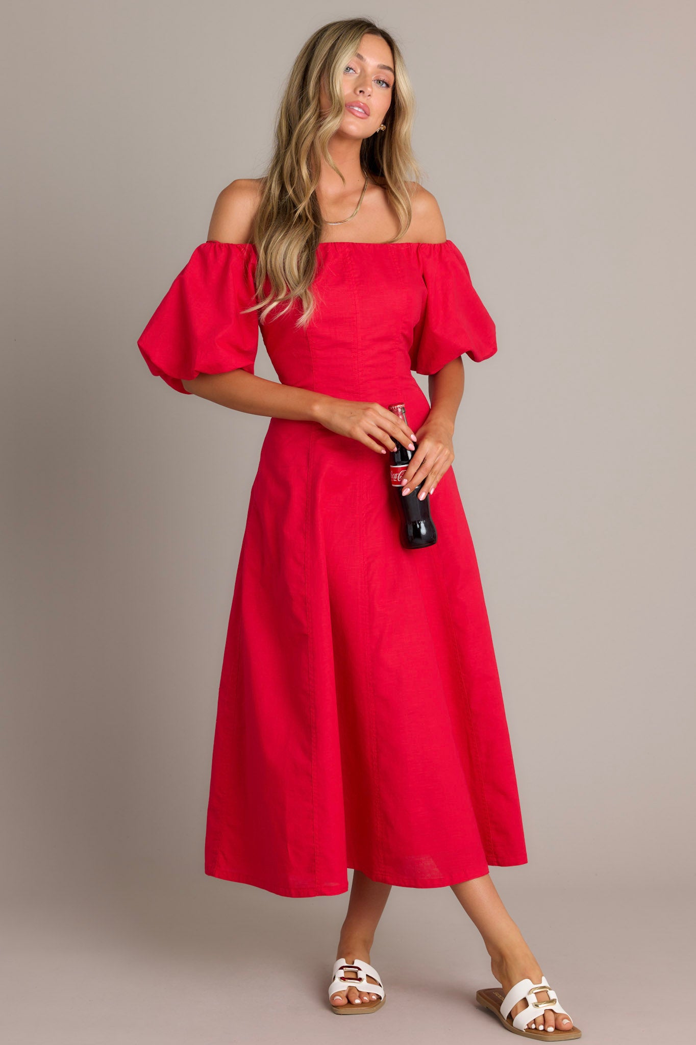 This red midi dress features an off shoulder elastic neckline, a discrete back zipper, functional hip pockets, visible front and back seams, elastic cuffed puff half sleeves, and a flowing silhouette.