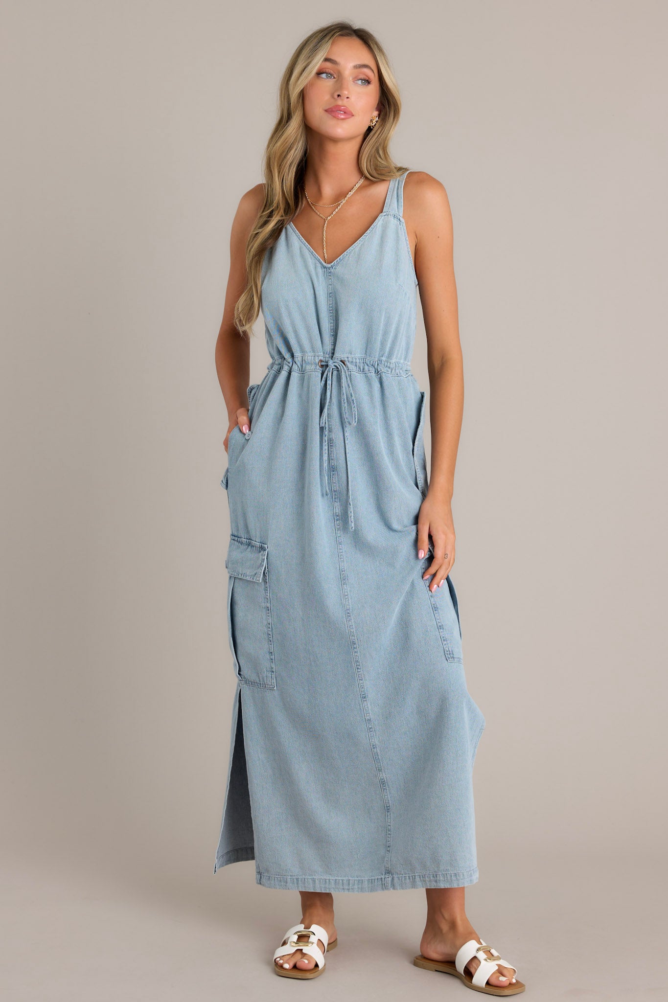 This chambray maxi dress features a v-neckline, thick adjustable straps, a self-tie drawstring waist, functional button hip, lower leg, and back pockets, and side slits.