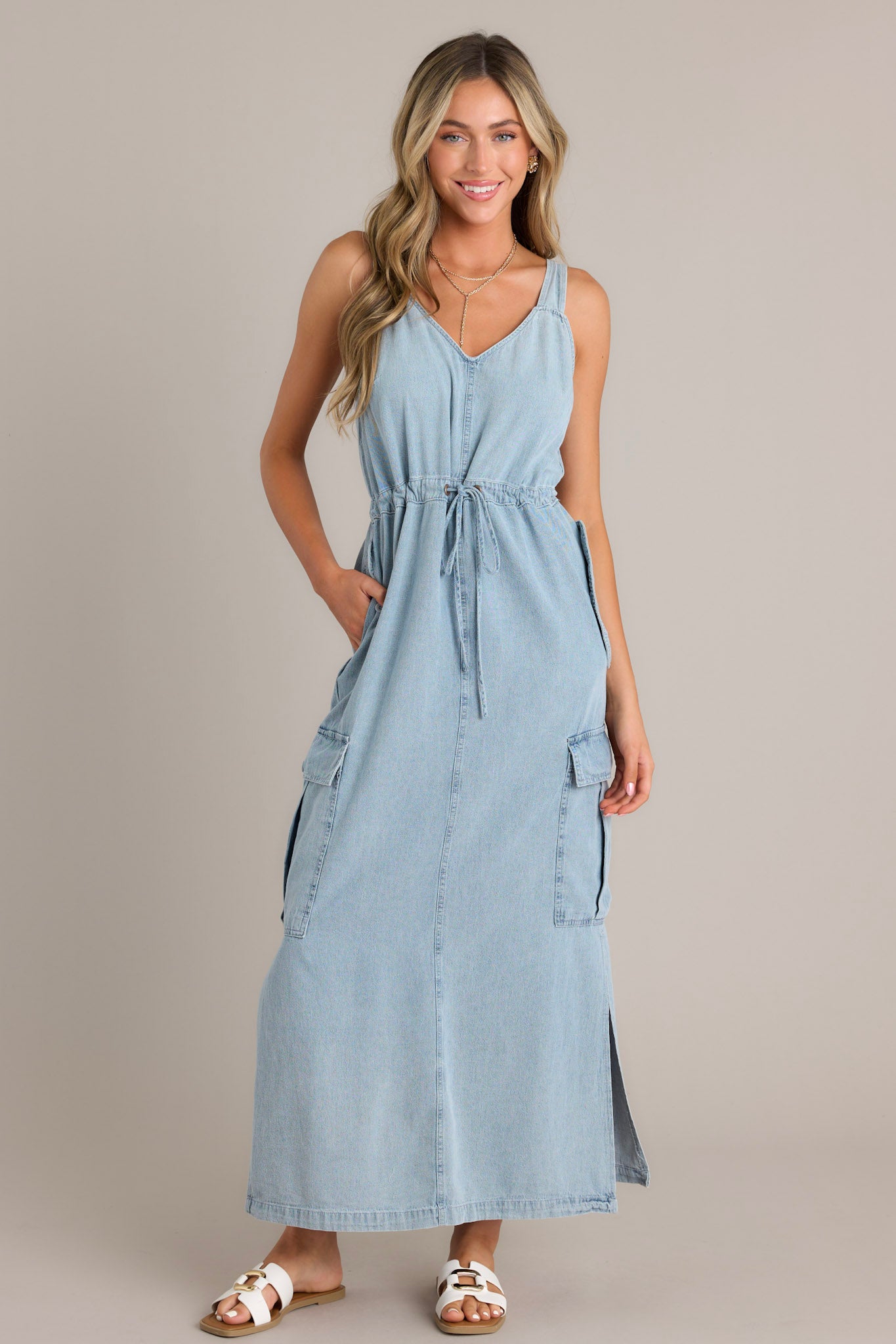 Front view of a chambray maxi dress featuring a v-neckline, thick adjustable straps, a self-tie drawstring waist, functional button hip, lower leg, and back pockets, and side slits.