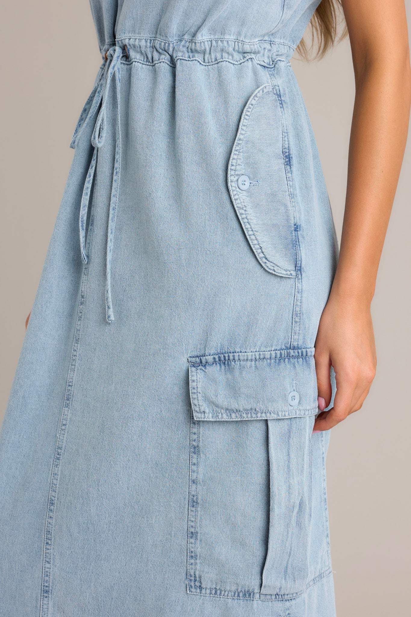 Close-up of the v-neckline, thick adjustable straps, self-tie drawstring waist, and functional button pockets of a chambray maxi dress.