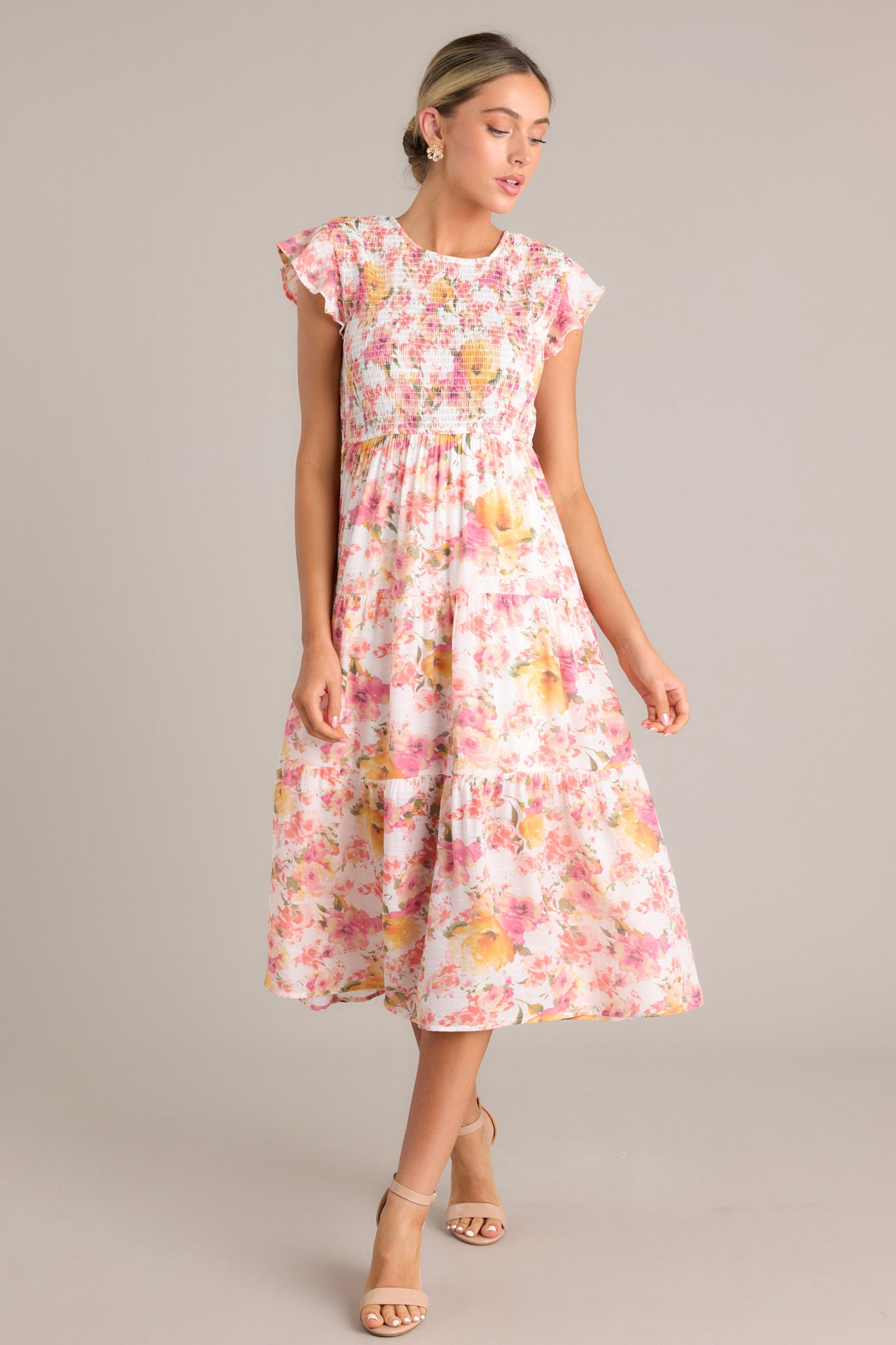 This pink midi dress features a high neckline, flutter sleeves, smocked bodice, tiered layers, and a lively floral design.