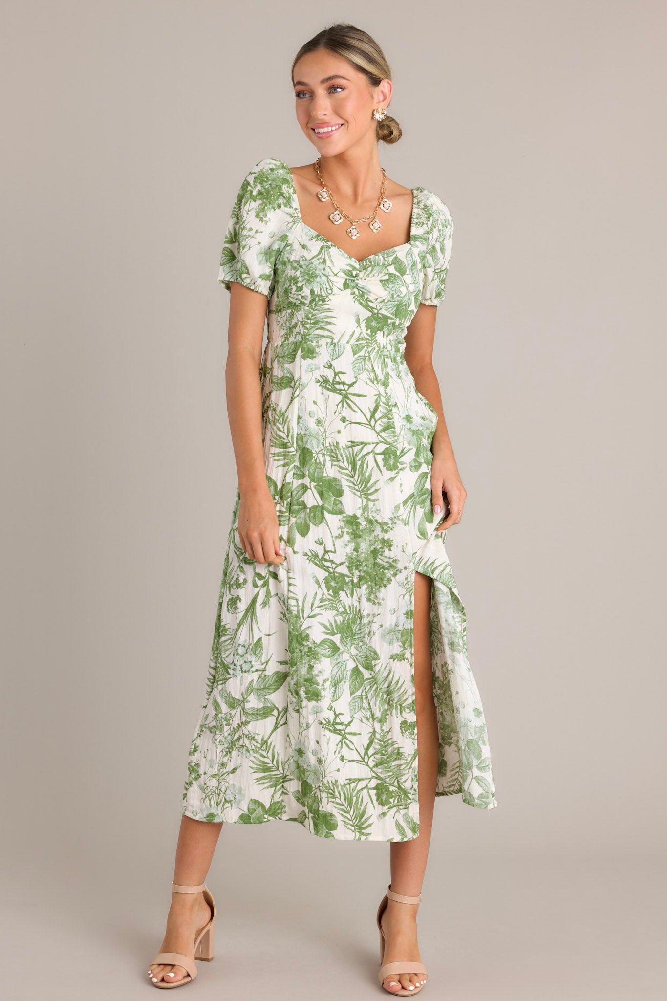 This midi dress features a green botanical print, short puff sleeves, a sweetheart neckline, fitted bodice, cinched waist, and a front slit on the mid-length skirt.