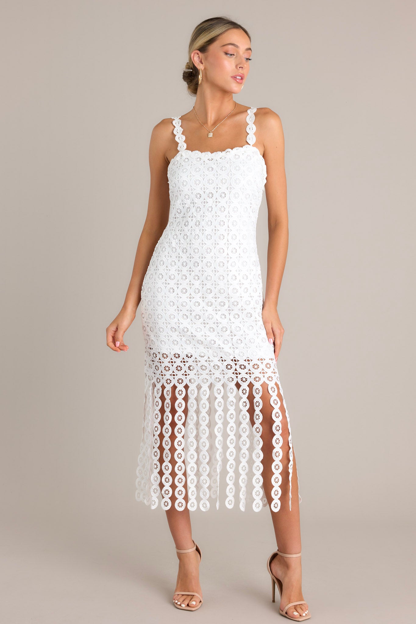 Front angled view of a white midi dress featuring a square neckline, detailed adjustable straps, a discrete side zipper, a unique lace pattern, and detailed hanging fringe