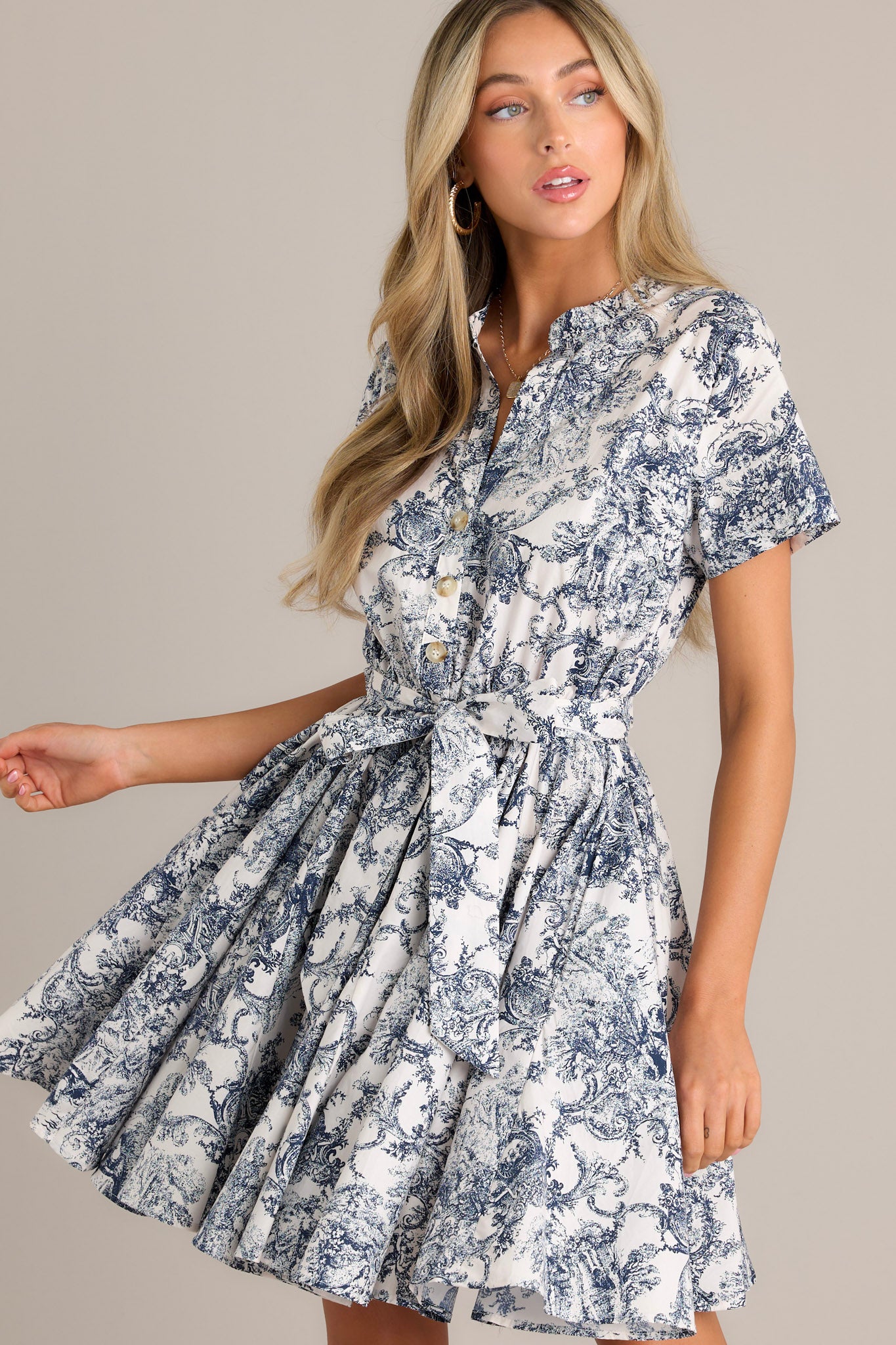 Front angled view of a navy mini dress featuring a v-neckline, button front bodice, elastic waistband, self-tie waist belt, belt loops, short sleeves, and a flowing silhouette.