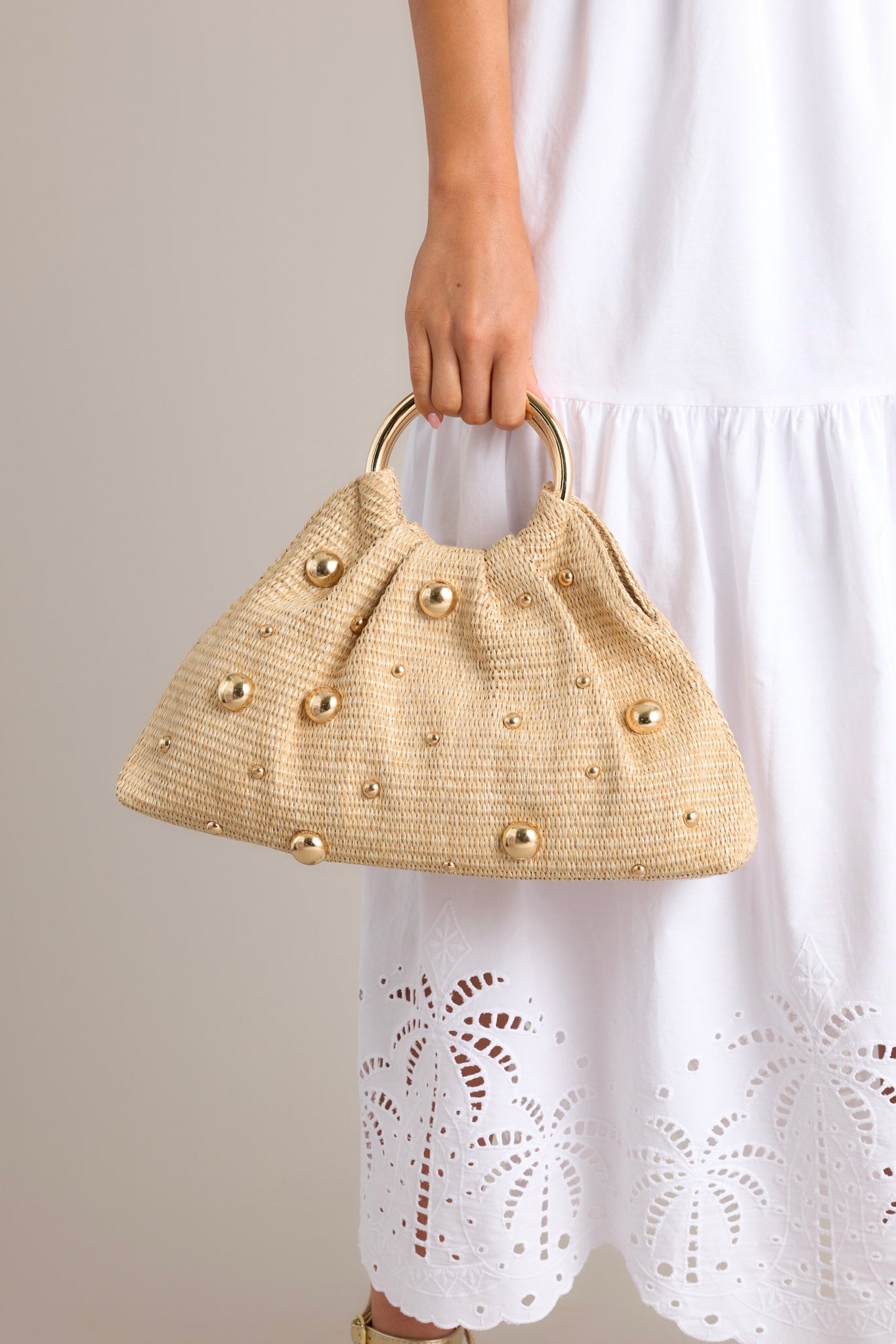 This rattan clutch features round shiny gold handles, a magnetic closure, a spacious interior with a smaller pocket inside, and round shiny gold embellishments that vary in size.