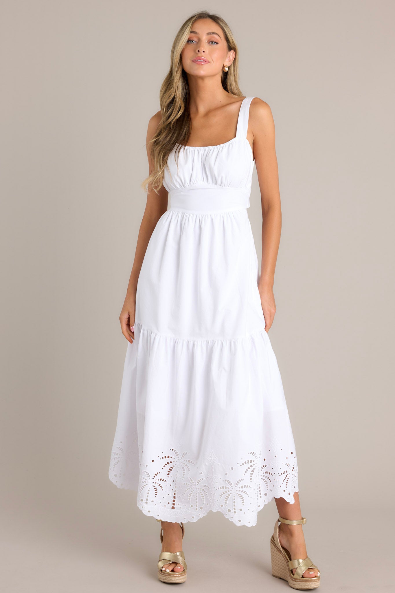 A woman wearing a white midi dress featuring an elastic square neckline, thick adjustable straps, a wide waistband with a self-tie back, an open lower back, a single tier, and palm eyelet detailing along the scalloped hemline.