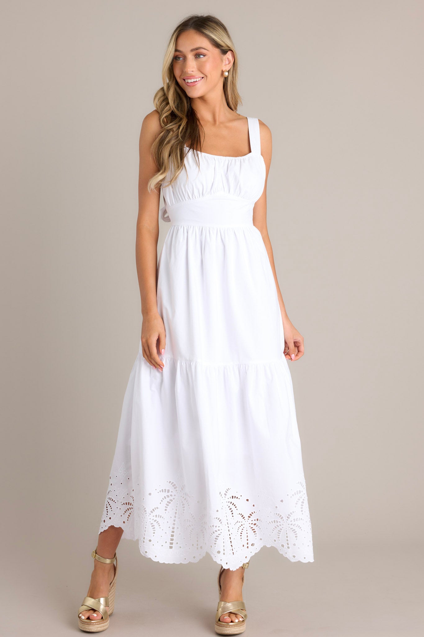 Full body view of a white midi dress with an elastic square neckline, thick adjustable straps, a wide waistband with a self-tie back, an open lower back, a single tier, and palm eyelet detailing along the scalloped hemline.