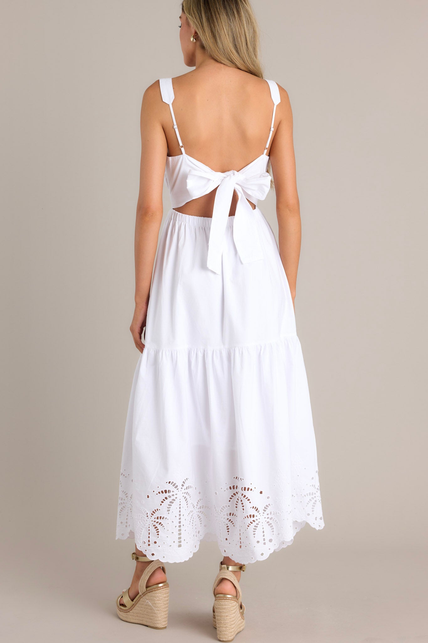 BAck view of a white midi dress with an elastic square neckline, thick adjustable straps, a wide waistband with a self-tie back, an open lower back, a single tier, and palm eyelet detailing along the scalloped hemline.