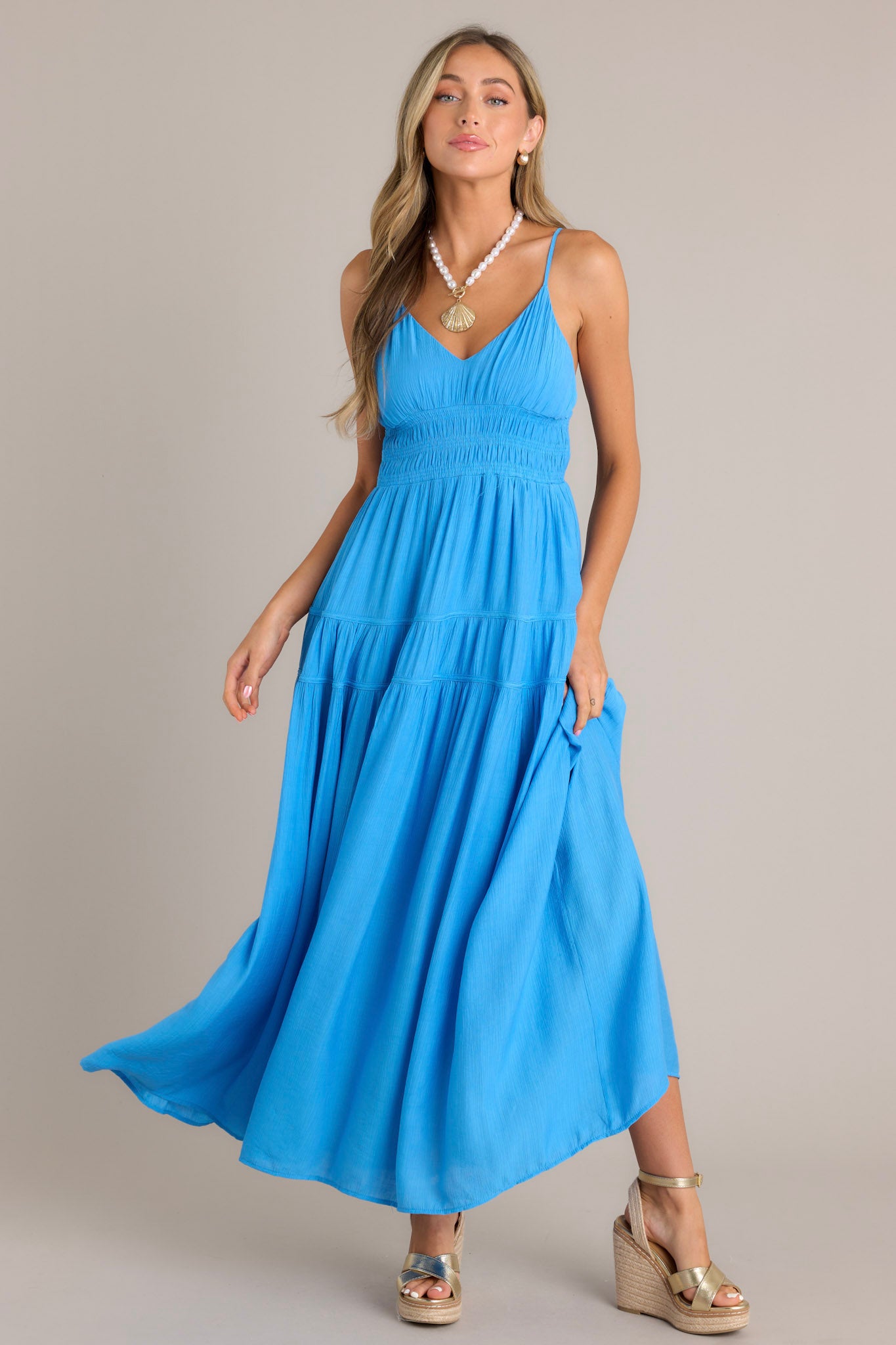 Front view of a blue maxi dress featuring a v-neckline, thin adjustable straps, a fully smocked waist, and a flowing tiered silhouette.