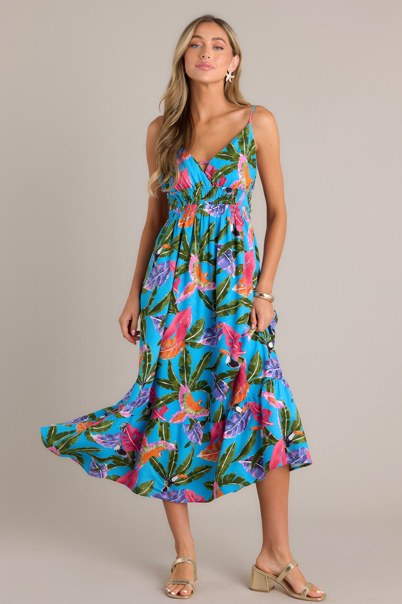 This blue midi dress features v-neckline, thin adjustable straps, an open back, a fully smocked waistband, a vibrant color scheme, and a flowing silhouette.