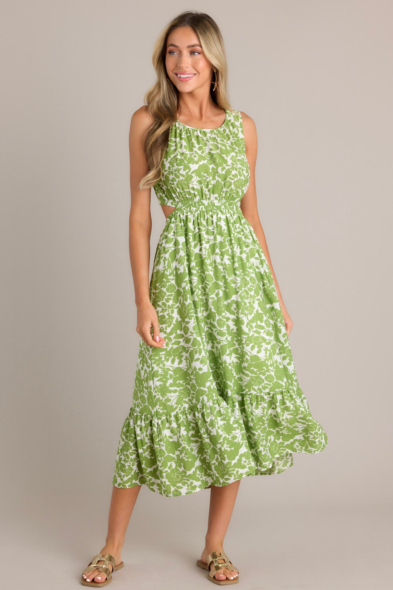 Action shot of a model wearing a green and white floral print sleeveless dress with side cutouts, a gathered waist, and a tiered skirt, highlighting the flow and movement of the dress.