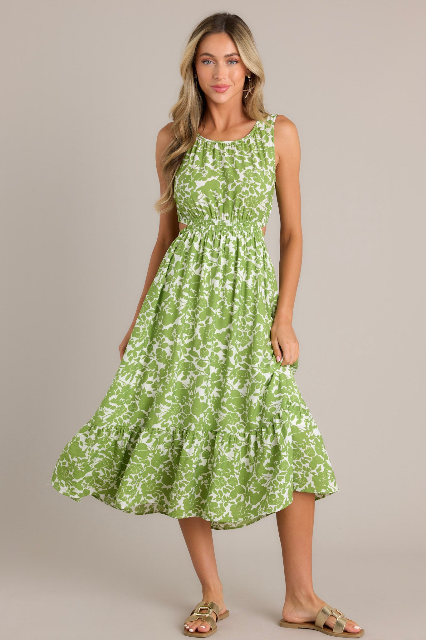 Front view of a model wearing a green and white floral print sleeveless dress with side cutouts, a gathered waist, and a tiered skirt.