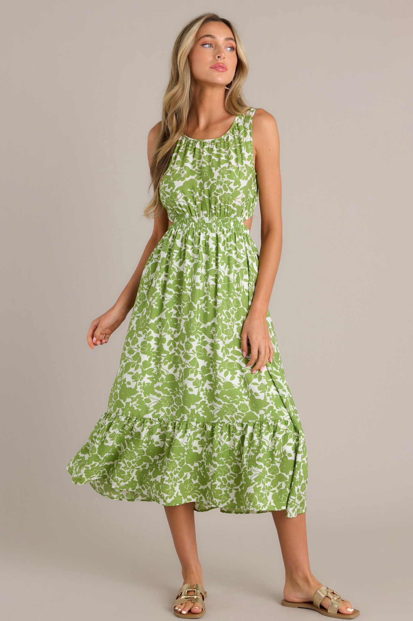Front angled view of a model wearing a green and white floral print sleeveless dress with side cutouts, a gathered waist, and a tiered skirt, showcasing the dress's flattering fit and design details.