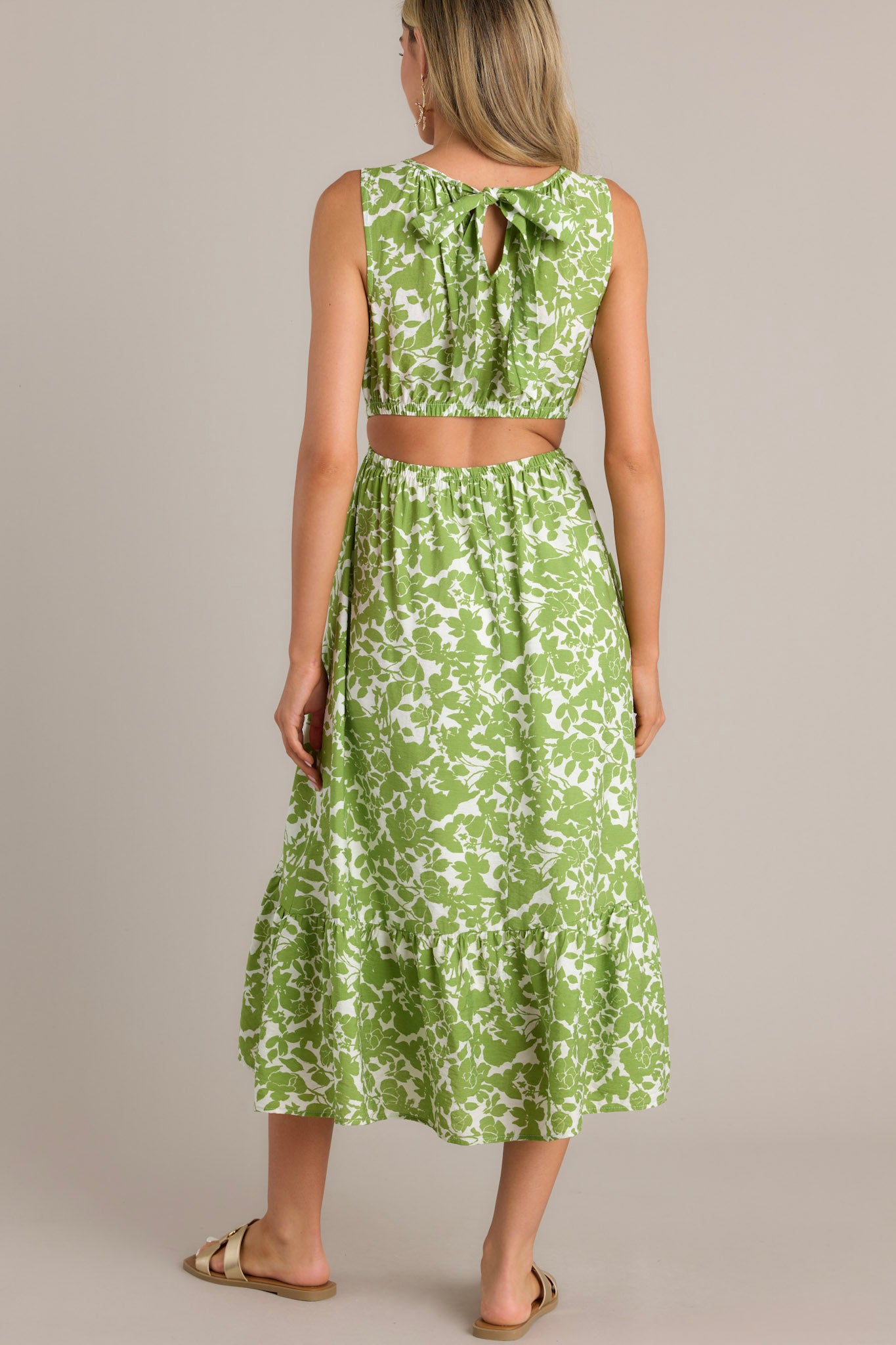 Back view of a green and white floral print sleeveless dress with a back bow detail, side cutouts, and a gathered waist, highlighting the open back and flowy tiered skirt.