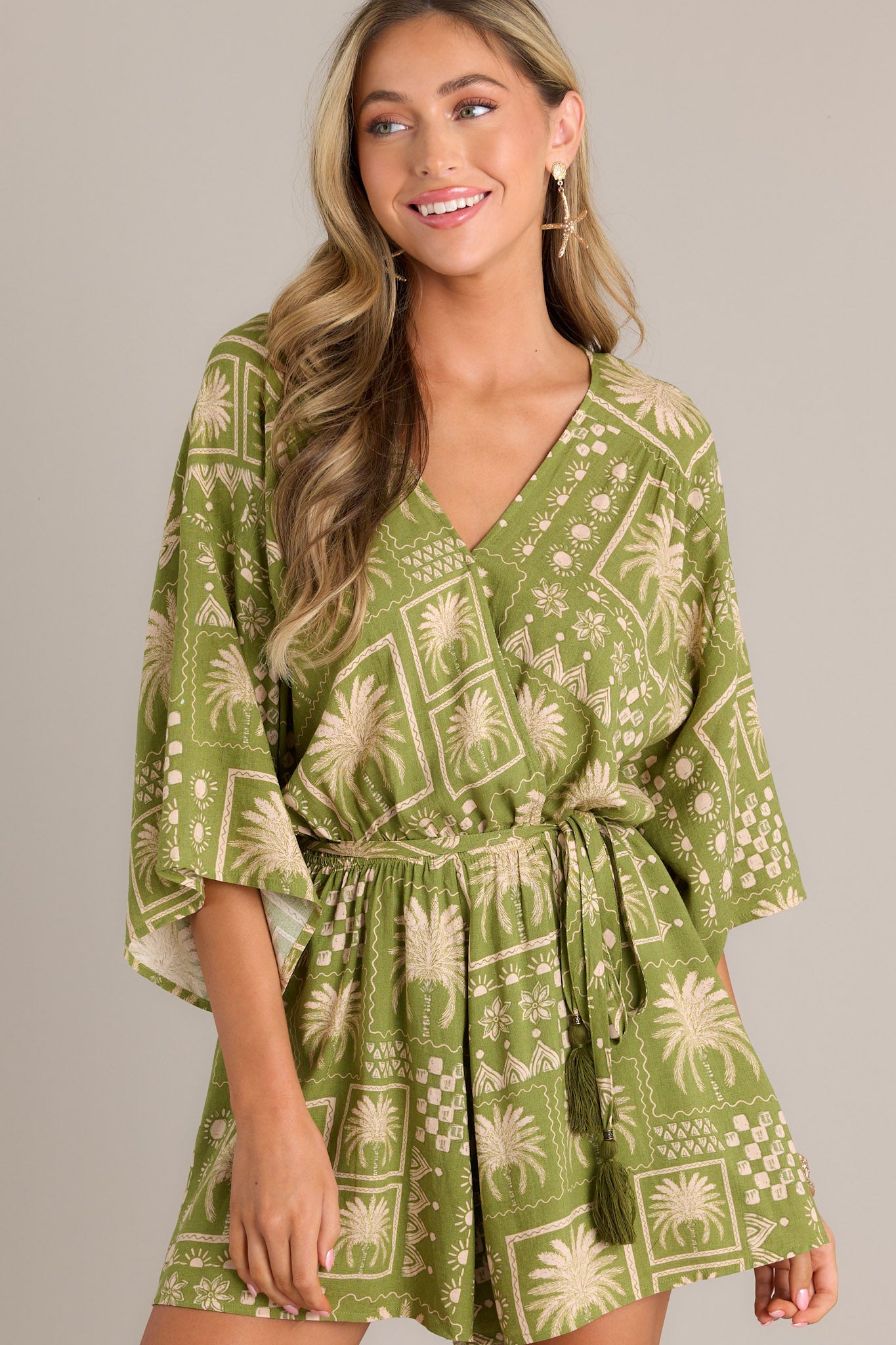 Front view of a green romper featuring a V-neckline, functional snap button closure, elastic waistband, self-tie belt, and quarter length dolman sleeves.