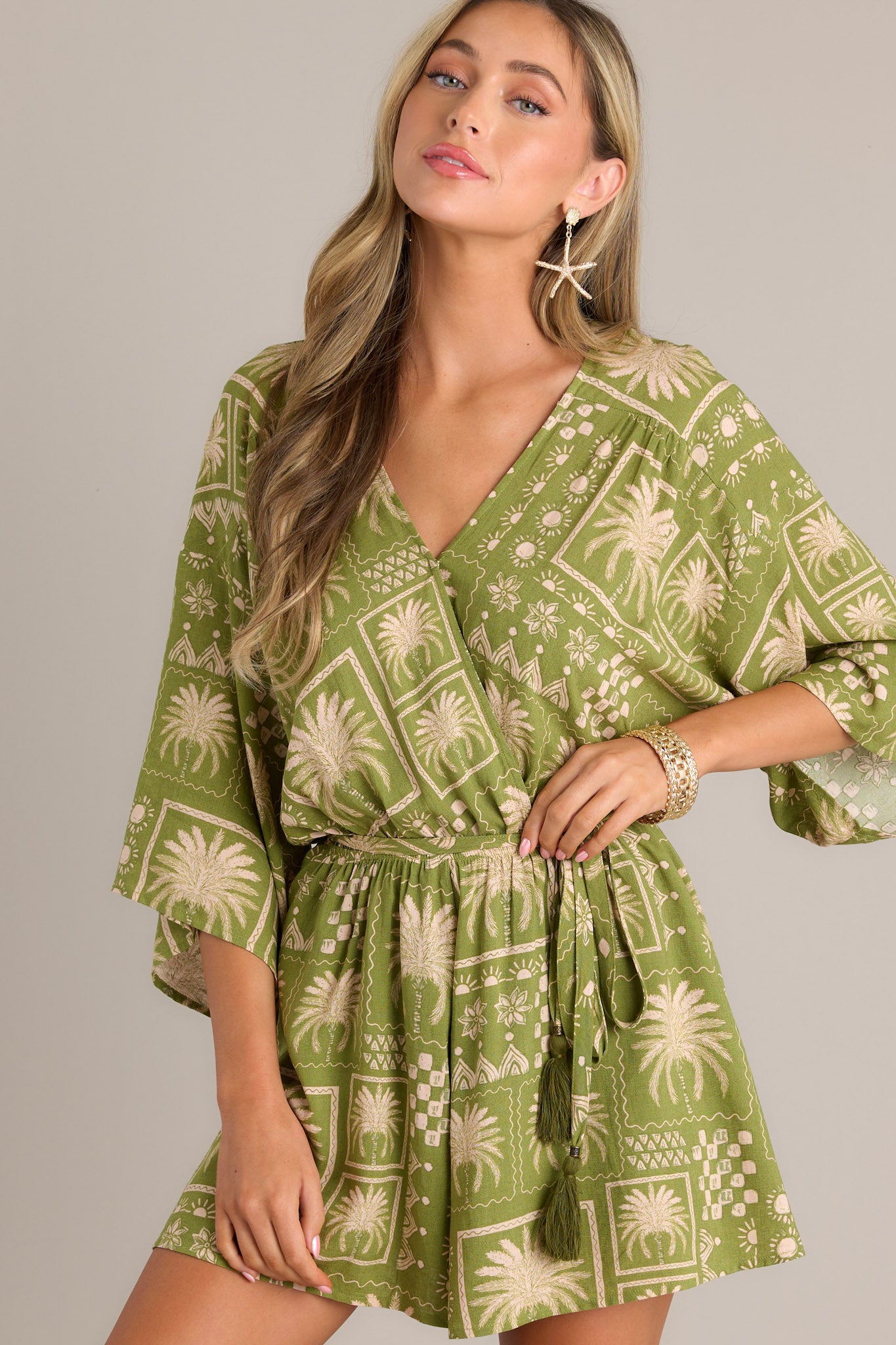 Angled Front view of a green romper featuring a V-neckline, functional snap button closure, elastic waistband, self-tie belt, and quarter length dolman sleeves.