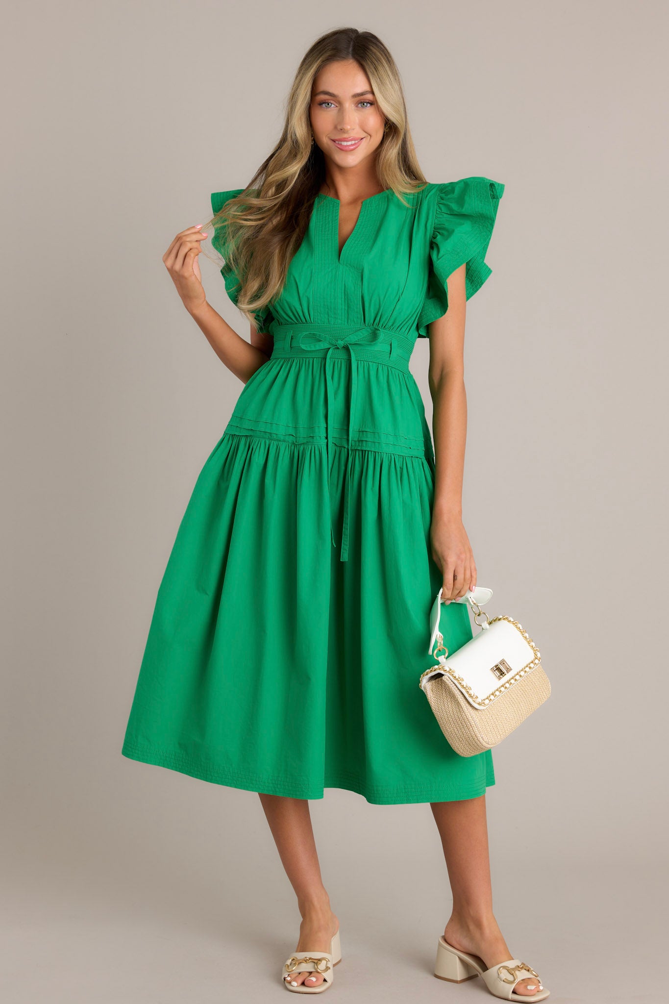 Full length view of a green midi dress with a v-neckline, subtle chest pleats, a discrete side zipper, a thick waistband with a self-tie belt, a smocked waist insert, functional hip pockets, a single tier, and ruffled short sleeves.