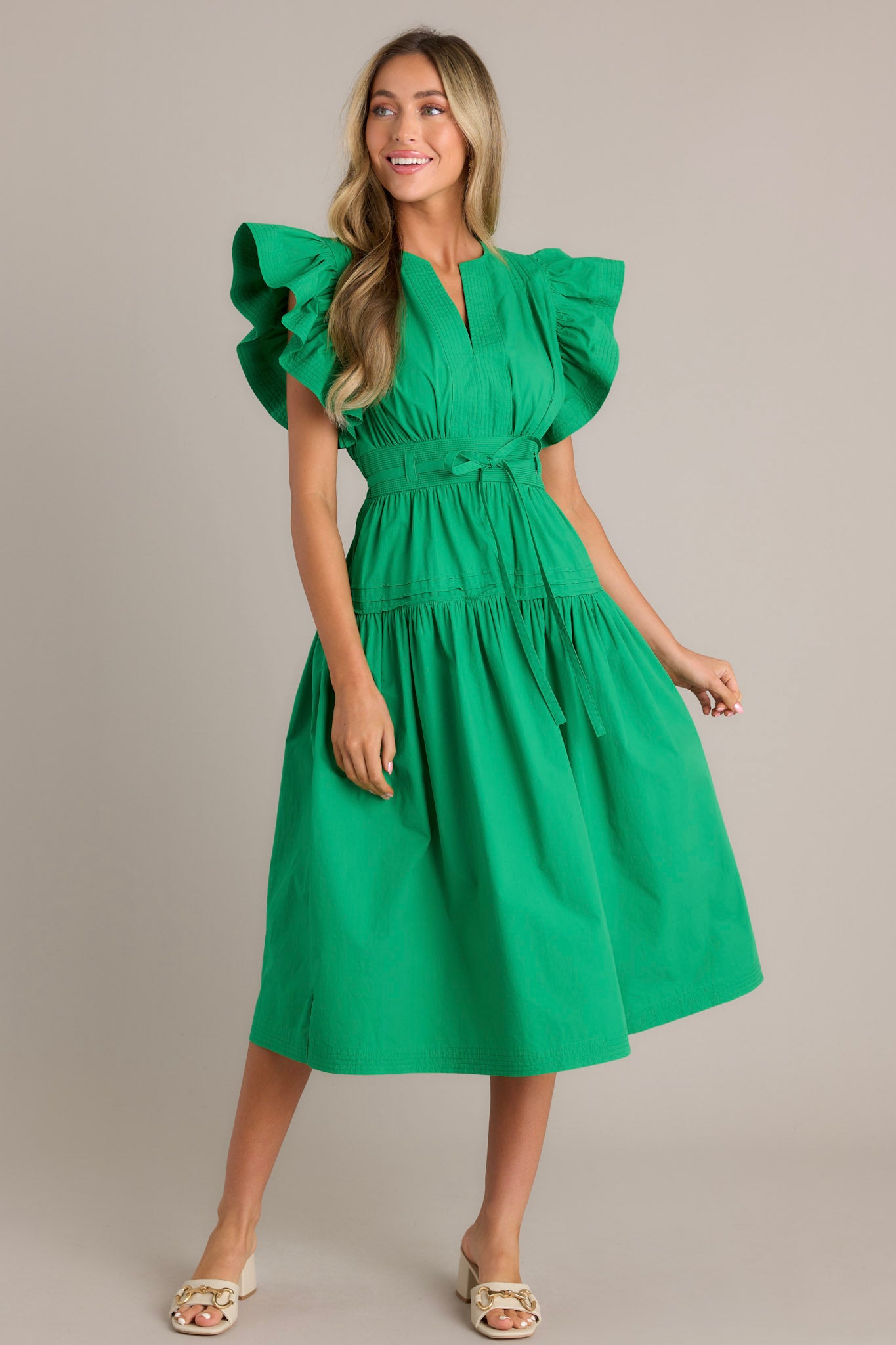 Action shot of a green midi dress highlighting the v-neckline, thick waistband with a self-tie belt, functional hip pockets, and ruffled short sleeves, showcasing the flow and movement of the dress.