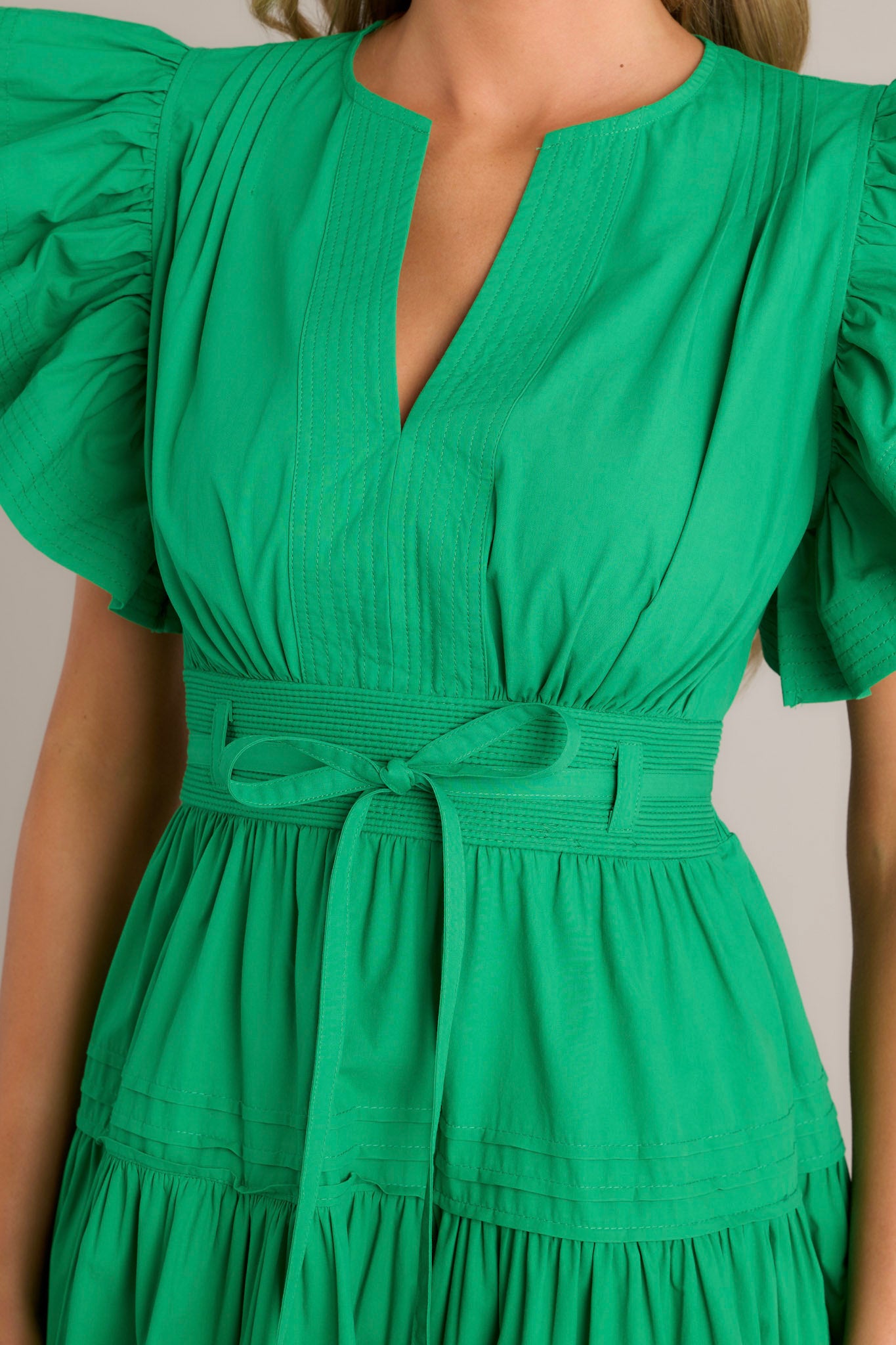 Close-up of the fabric and design details on a green midi dress featuring a v-neckline, subtle chest pleats, and a thick waistband with a self-tie belt.