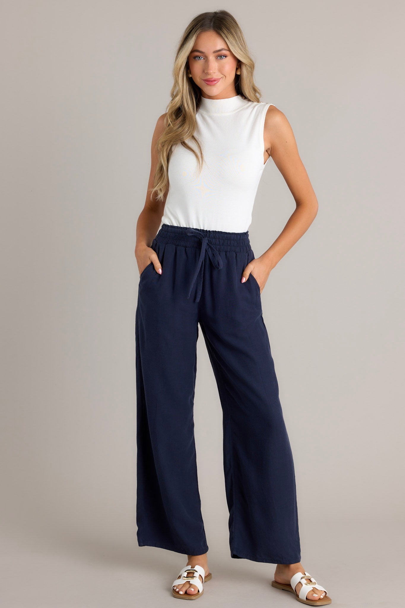 Angled full body view of the material of these navy pants that feature a high waisted design, an elastic waistband with a self-tie drawstring, functional front & back pockets, and a wide leg.