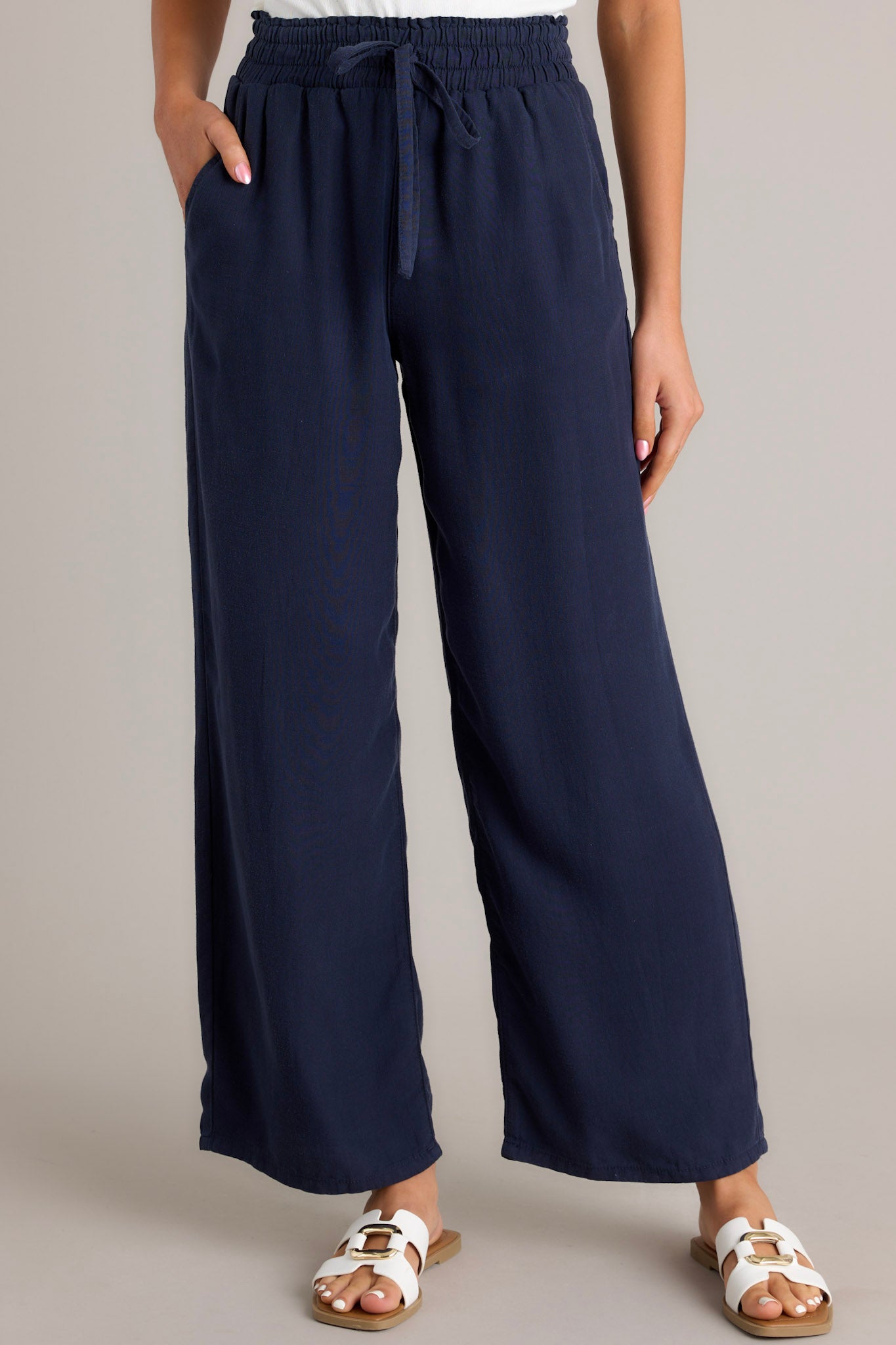 Front view of the material of these navy pants that feature a high waisted design, an elastic waistband with a self-tie drawstring, functional front & back pockets, and a wide leg.