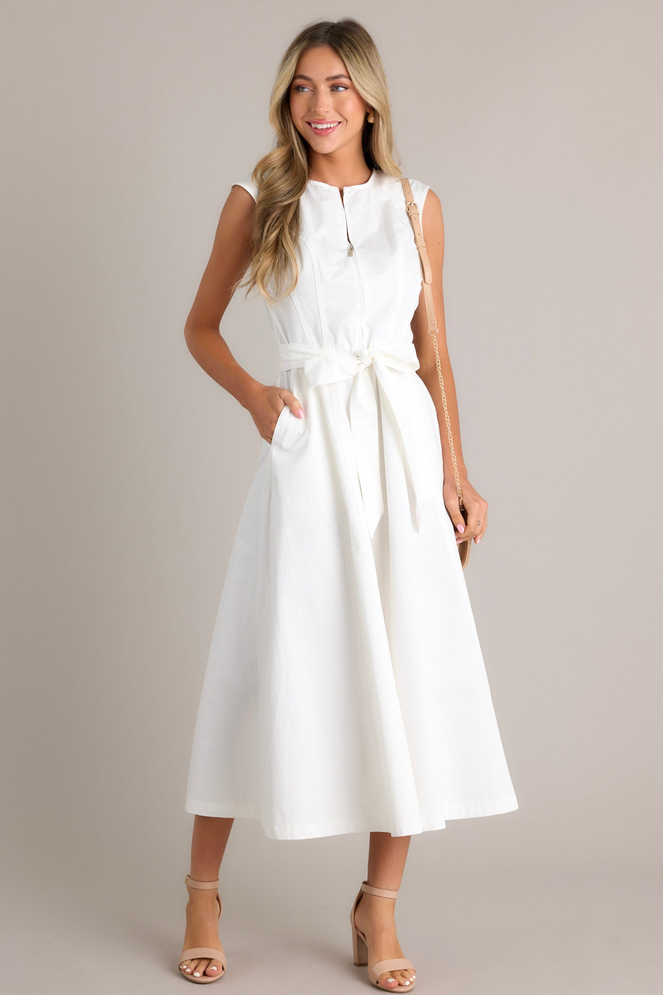 Full body view of this white midi dress that features a round neckline, a zipper front, visible front & back seams, a self-tie waist belt, belt loops, functional hip pockets, and a flowing silhouette.