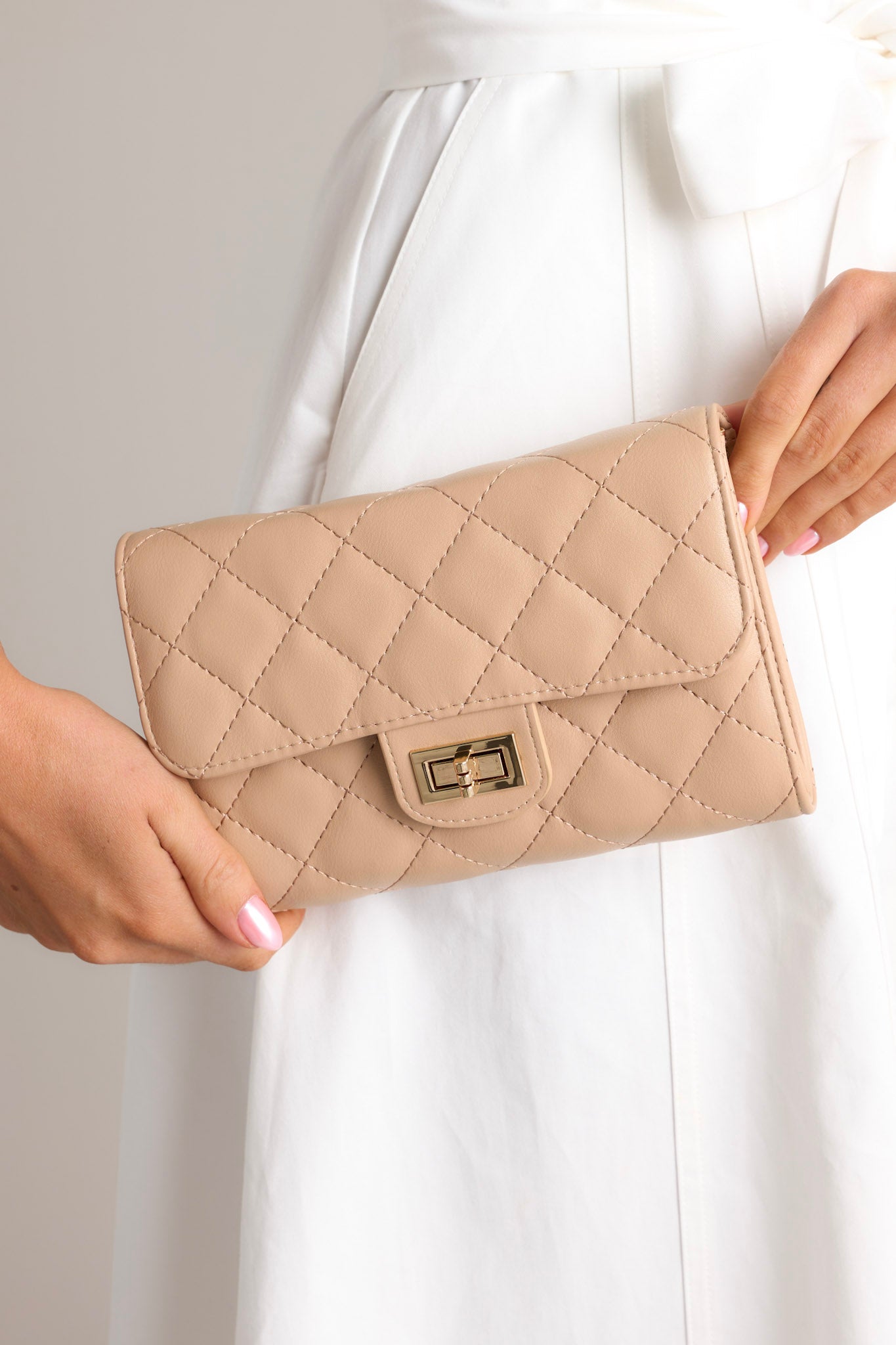 Close-up of a beige quilted clutch bag with a front turn-lock closure, held against a white dress