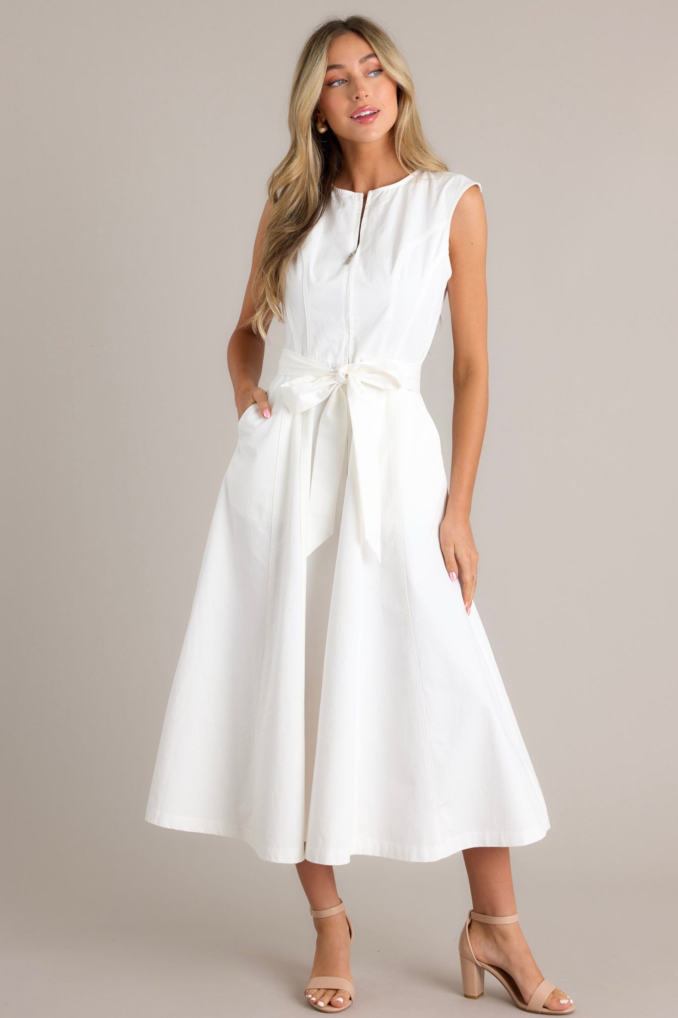 A white sleeveless midi dress with a front zipper, a fitted bodice, and a wide waist tie that forms a bow. The dress features a flared skirt and side pockets